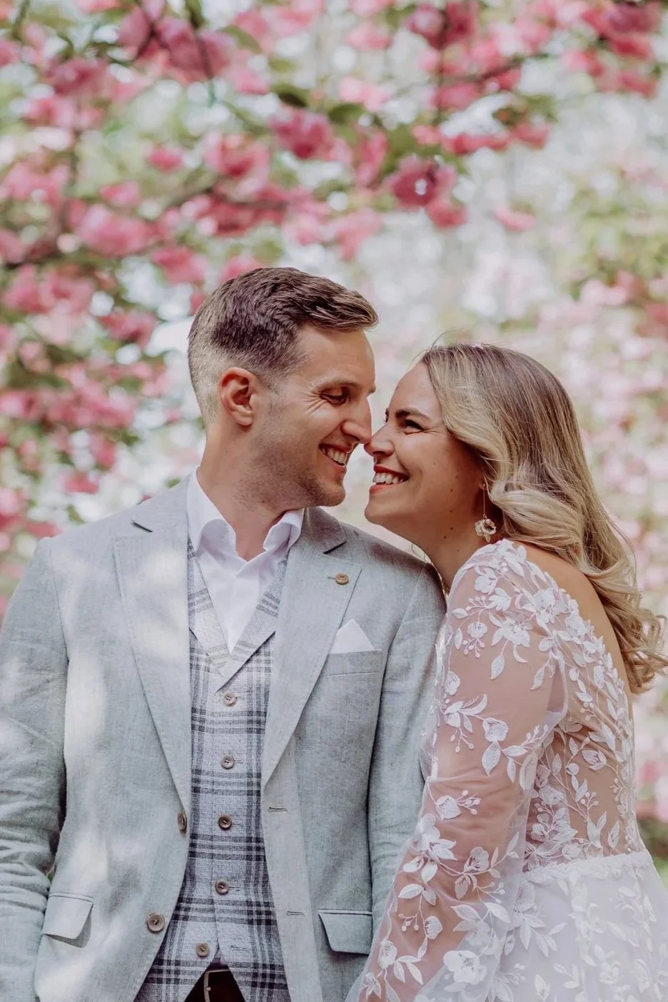 Ein glückliches Paar bei einer Hochzeit im Garten, umgeben von pinken Blüten, die sich lächelnd berühren.