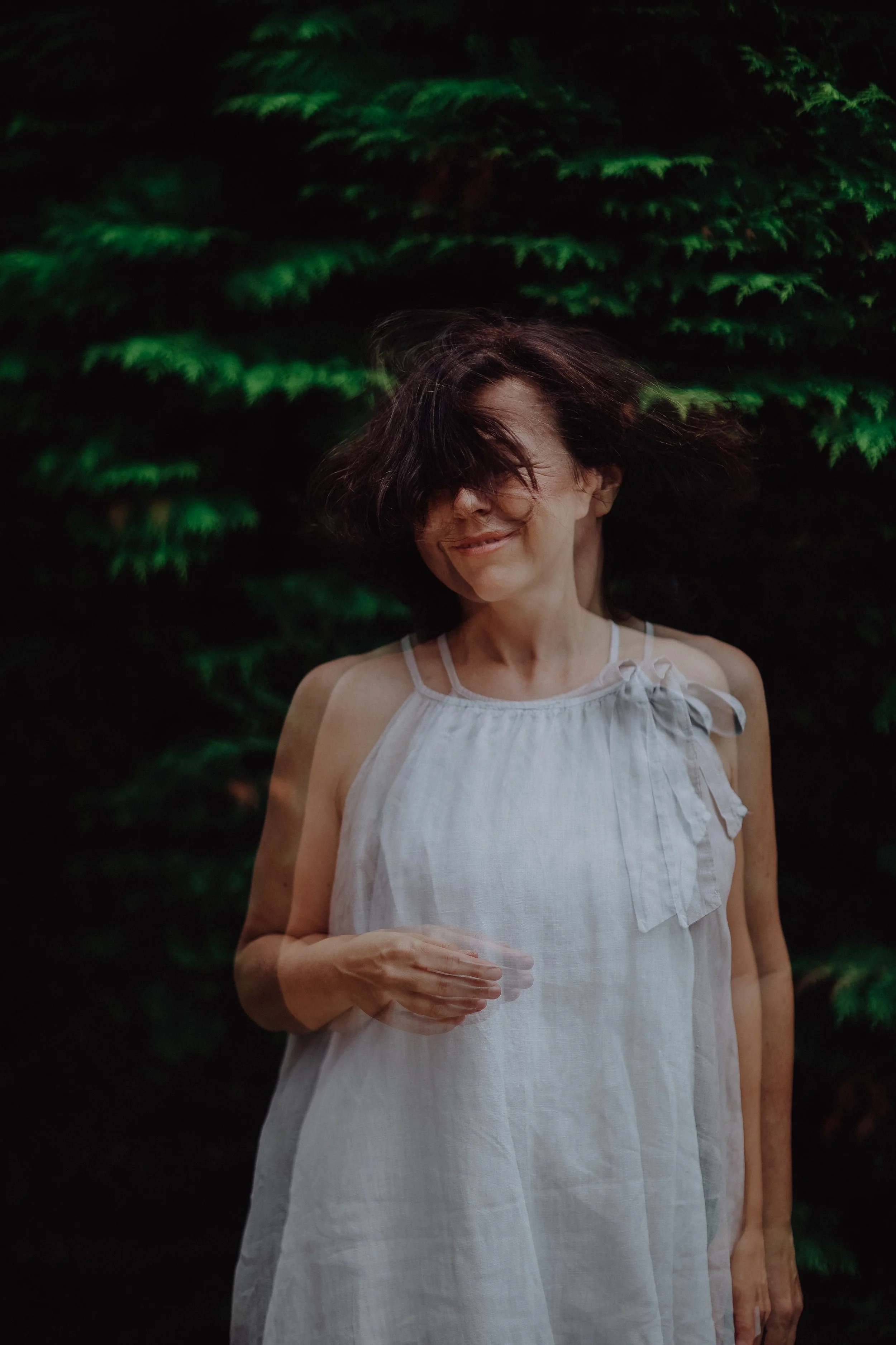 Eine lächelnde Frau in einem weißen Kleid vor grünen Büschen, mit Bewegungsunschärfe durch ihre Kopf- und Armbewegung.