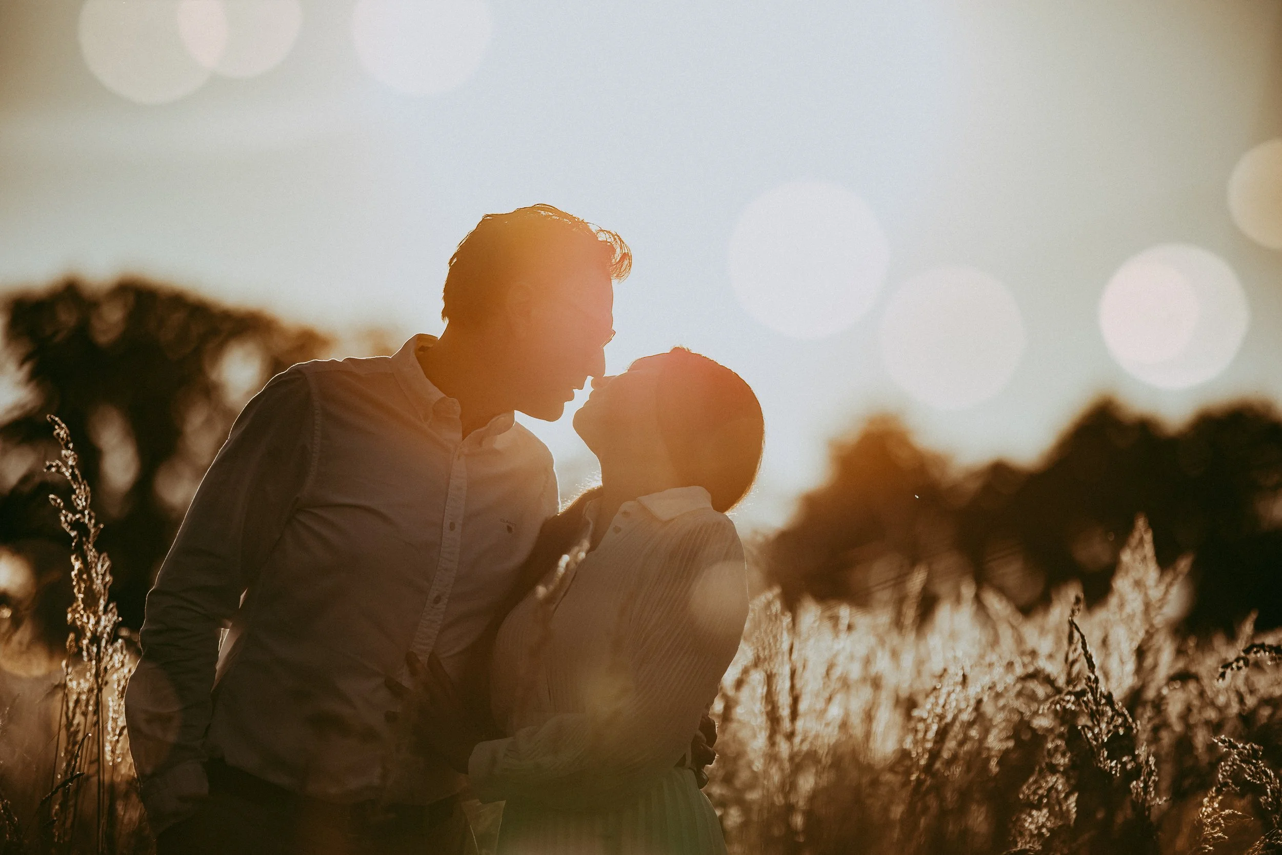 Ein Paar küsst sich im Sonnenuntergang in einem Feld, umgeben von Gräsern und Bäumen im Hintergrund.