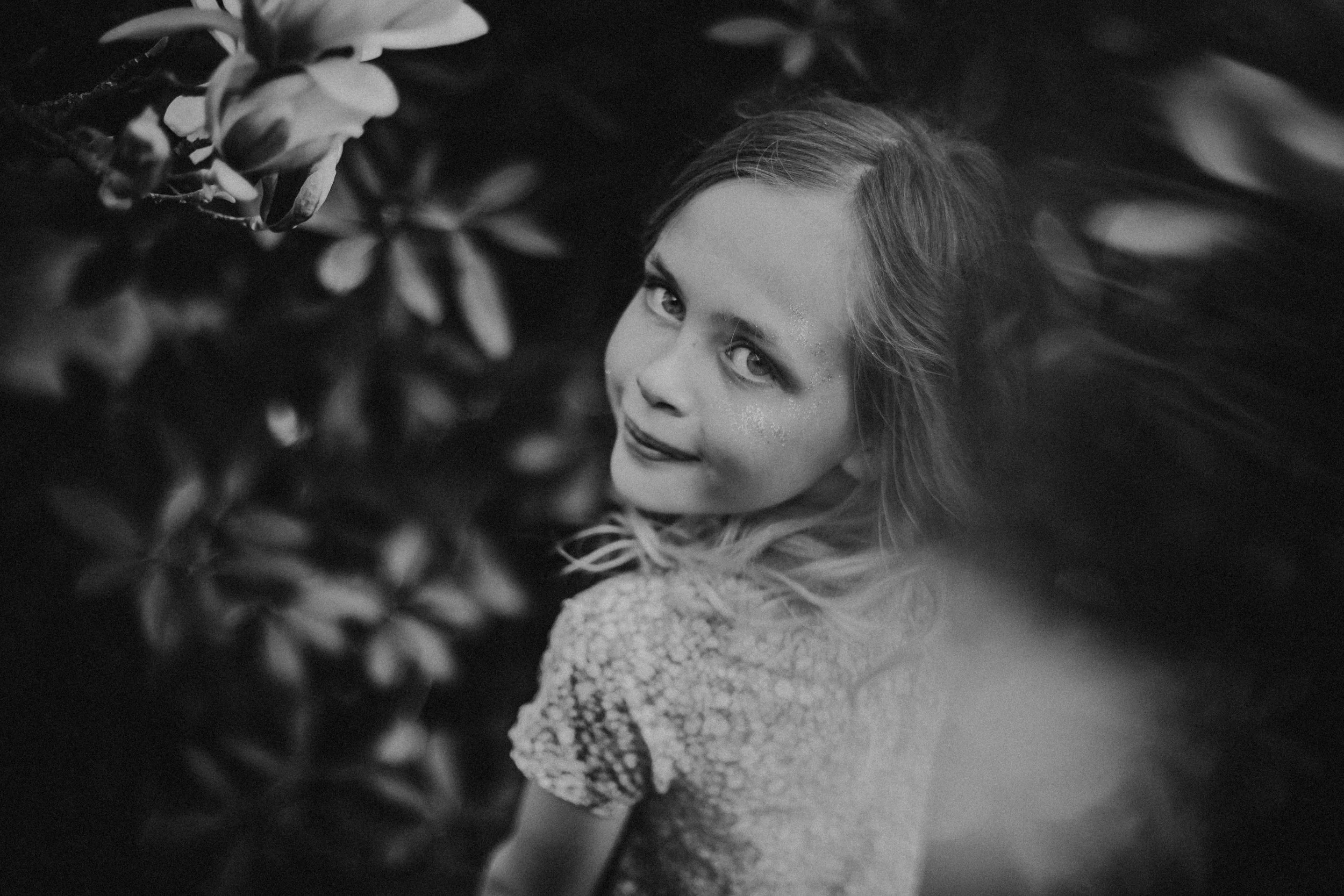 Ein junges Mädchen mit lockigem Haar und einem freundlichen Blick, umgeben von Blättern und Pflanzen, in Schwarz-Weiß aufgenommen.