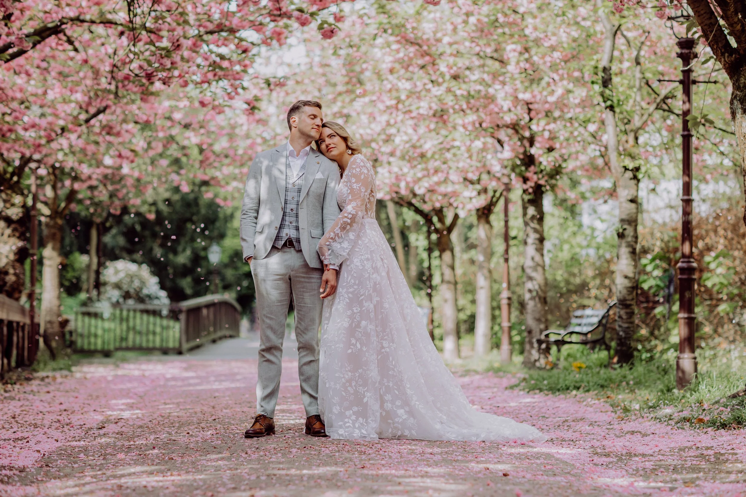 Ein Brautpaar steht auf einem Weg im Park, umgeben von blühenden Kirschbäumen.
