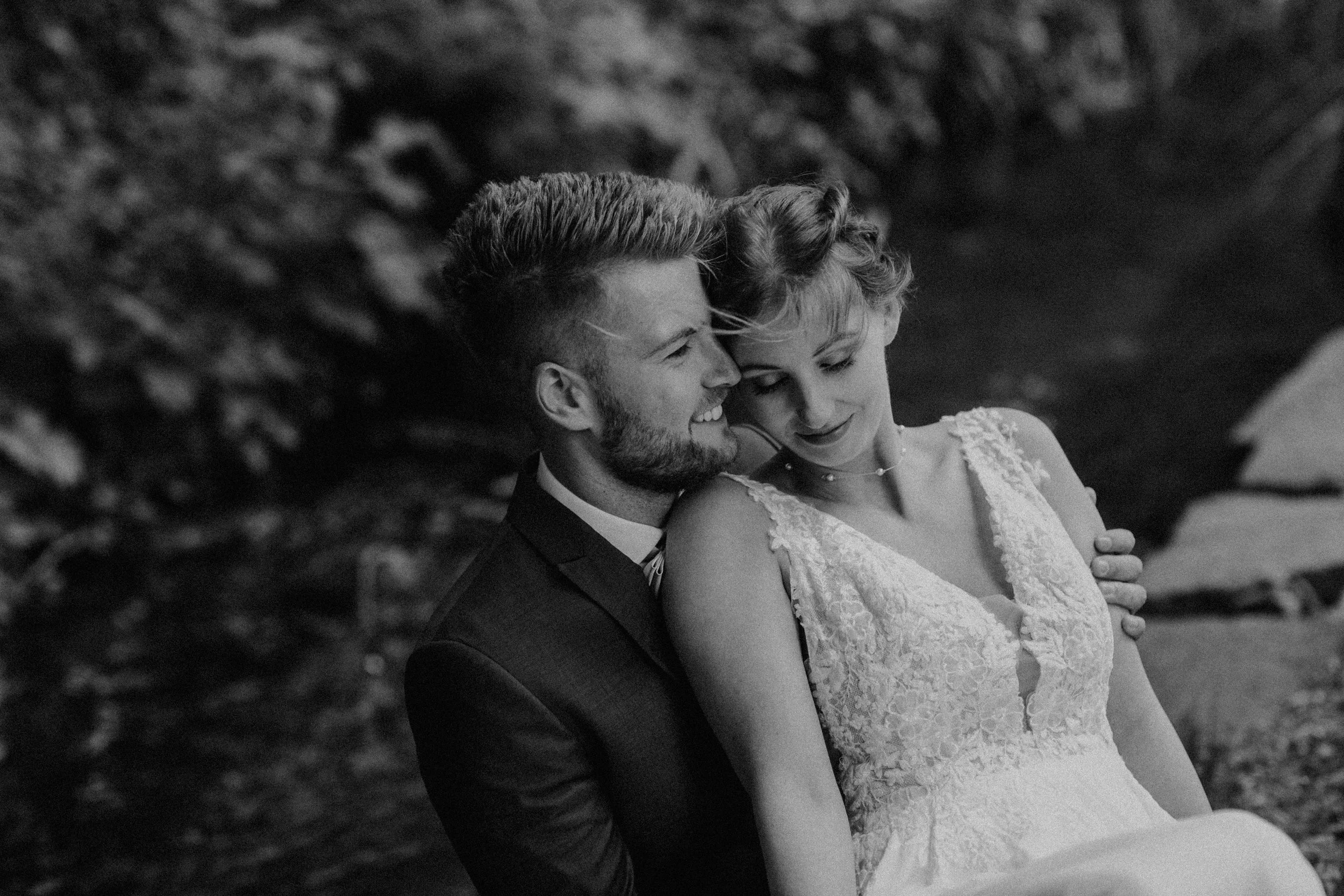 Braut und Bräutigam sitzen nahe beieinander draußen, lächeln und schauen liebevoll aufeinander, in Schwarz-Weiß-Fotografie.