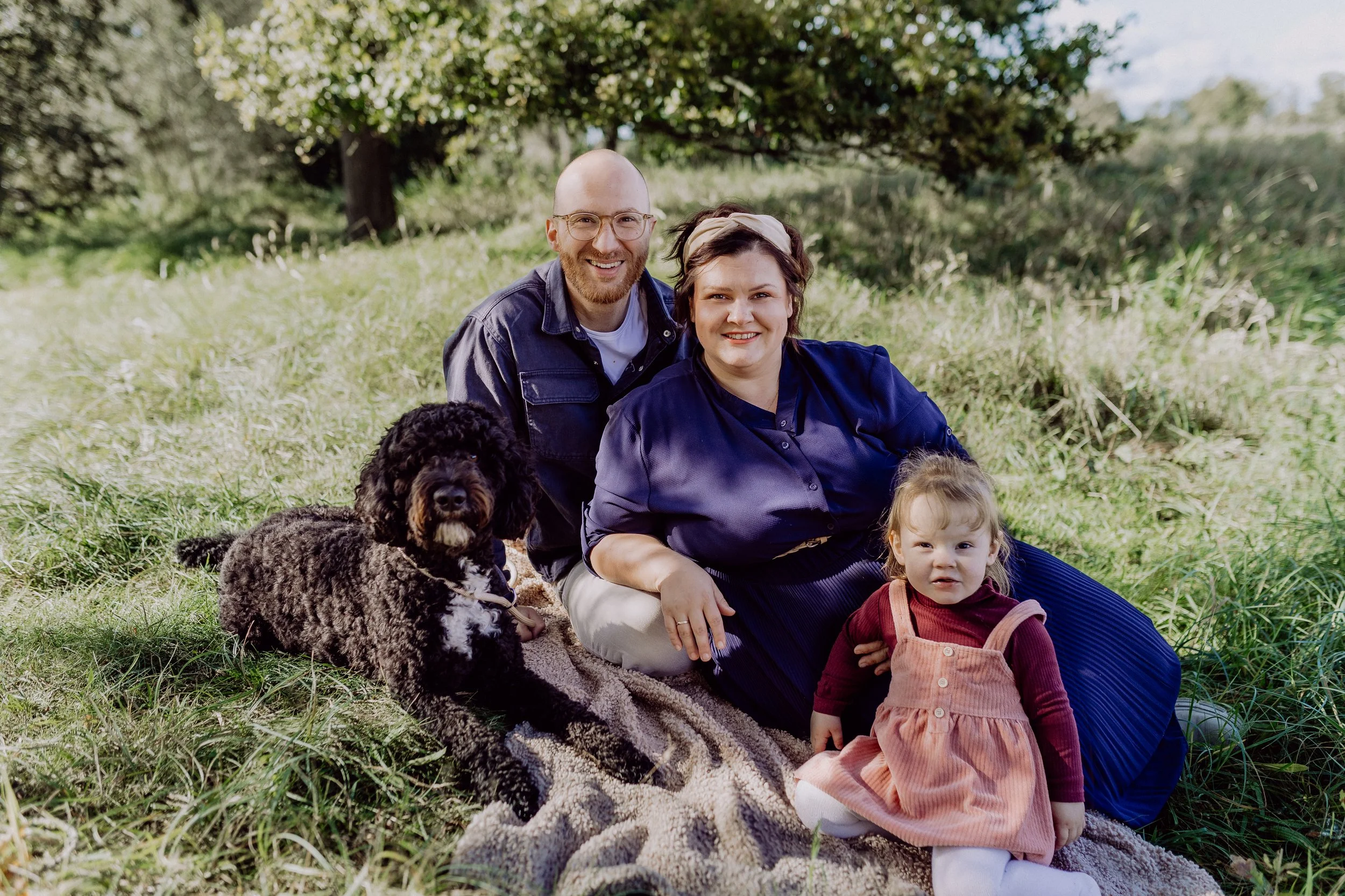 Familie mit einem Hund und einem kleinen Kind sitzt auf einem Decke im Grünen, umgeben von Bäumen und Gras, bei schönem Wetter.