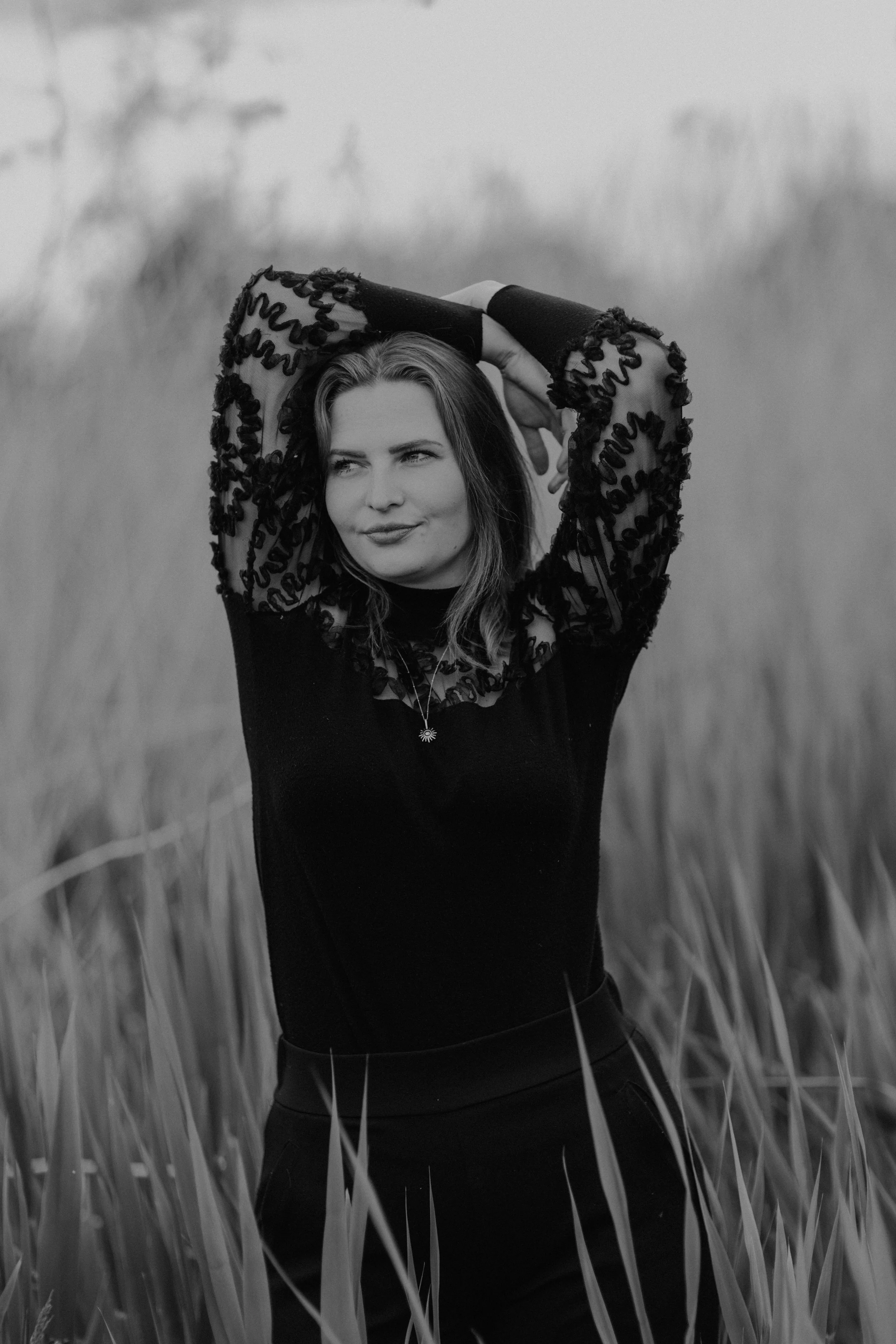 Schwarz-weiße Porträtaufnahme einer jungen Frau mit lockigem Haar, die in einem Feld steht. Sie trägt ein elegantes Top mit gesticktem Ärmel-Design und eine Halskette, schaut leicht seitlich und lächelt sanft.