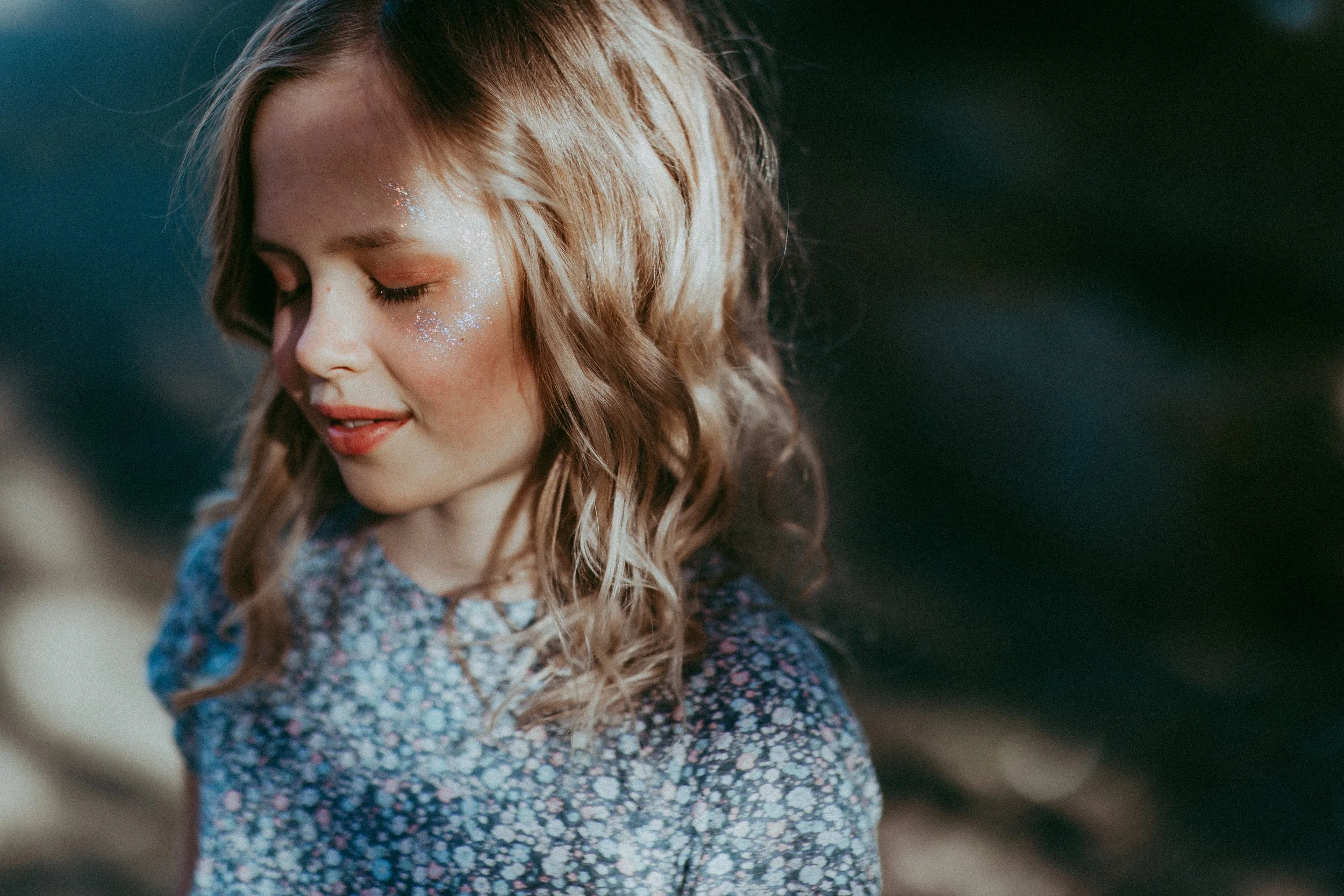 Ein junges Mädchen mit lockigem, hellbraunem Haar, das nach einem Sonnenbad mit Glitzer auf ihrem Gesicht lächelt, trägt ein bunten, floral bedrucktes Kleid vor einem unscharfen Hintergrund im Freien.