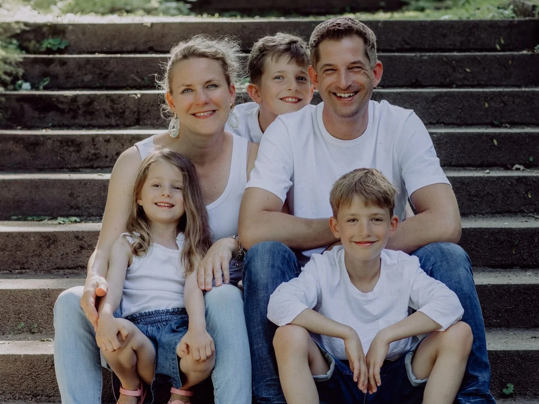 Eine lächelnde Familie mit vier Mitgliedern sitzt auf Steinstufen im Freien, alle sind in weißen Oberteilen gekleidet.