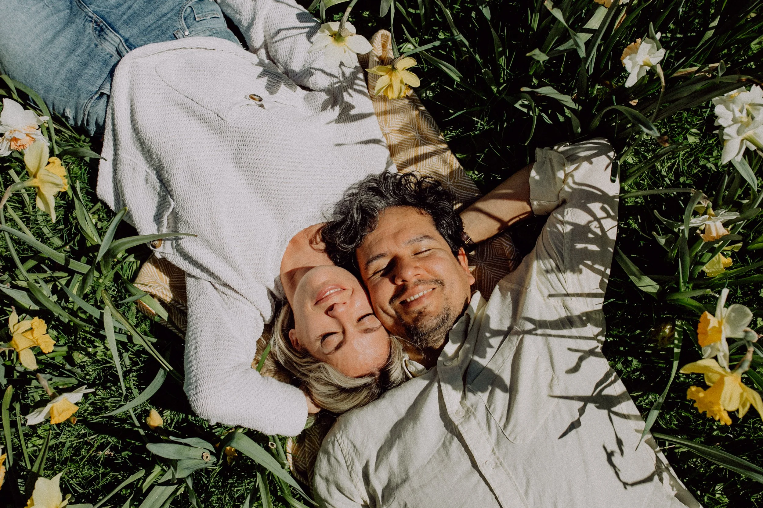 Ein lachendes Paar liegt auf einer Decke im Garten, umgeben von gelben und weißen Blumen, bei Sonnenschein.