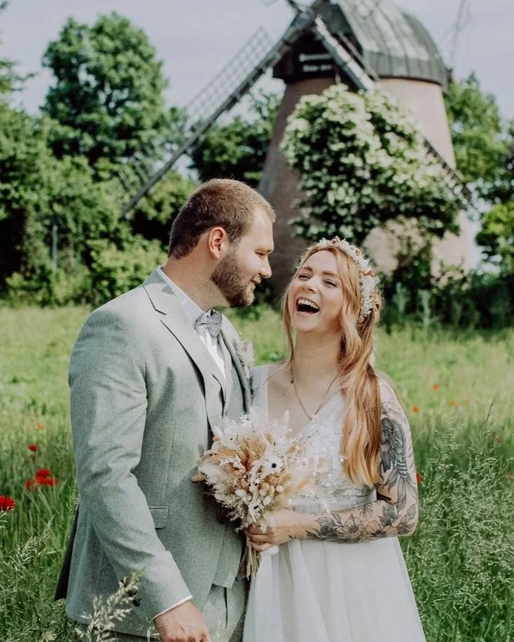 Ein brautpaar mit einem Mann in einem grauen Anzug und einer Frau mit langen roten Haaren in einem weißen Brautkleid stehen im Grünen vor einer Windmühle, lachen und halten einen Brautstrauß.