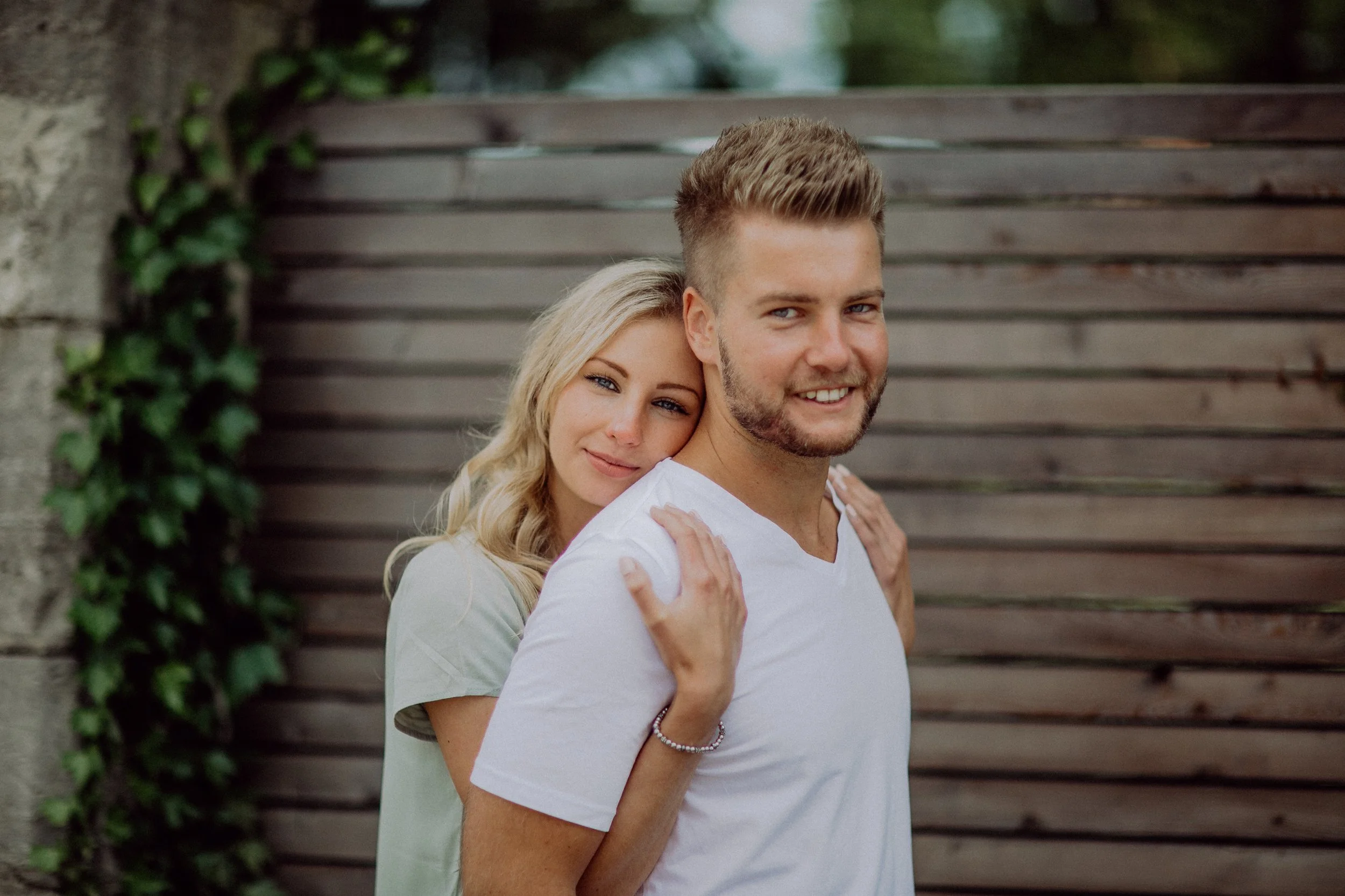 Ein lächelndes Paar steht vor einer Holzlaube, die Frau liegt auf den Schultern des Mannes.