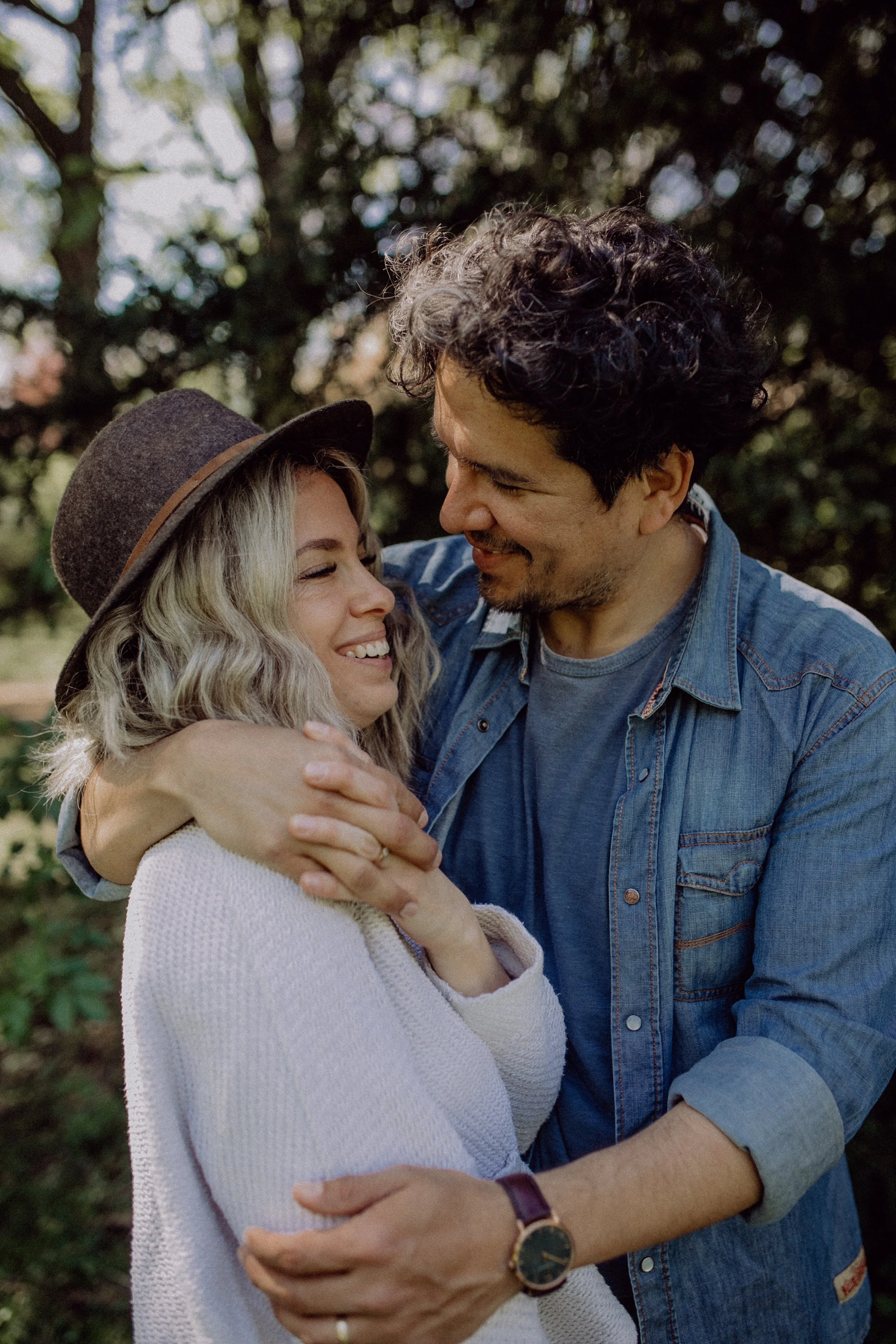 Ein Mann und eine Frau umarmen sich im Freien, lächeln sich liebevoll an, im Hintergrund Bäume und natürliches Licht.