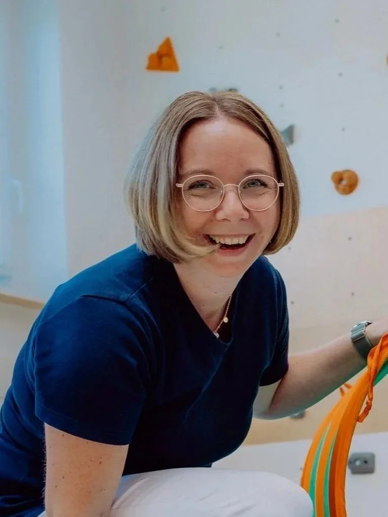 Eine lachende Frau mit Brille und blonden, schulterlangen Haaren, sitzt neben einem Kletterwand mit bunten Griffen.