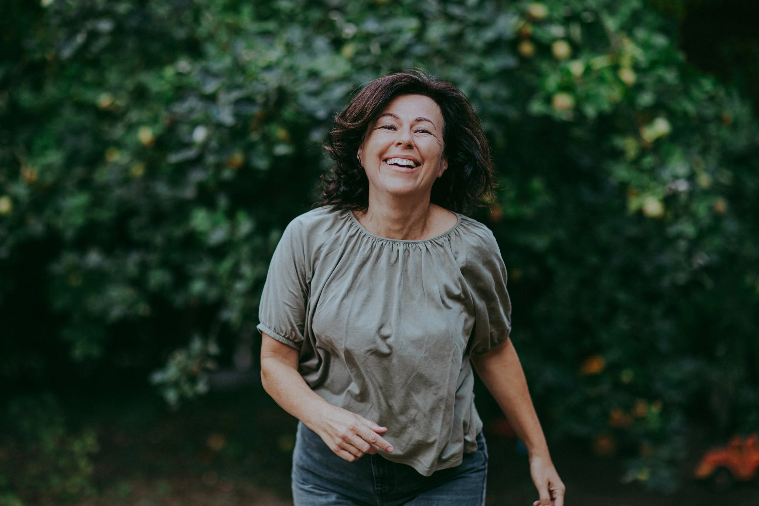 Eine fröhliche Frau mit dunklen, gewellten Haaren, die im Grünen draußen lacht.