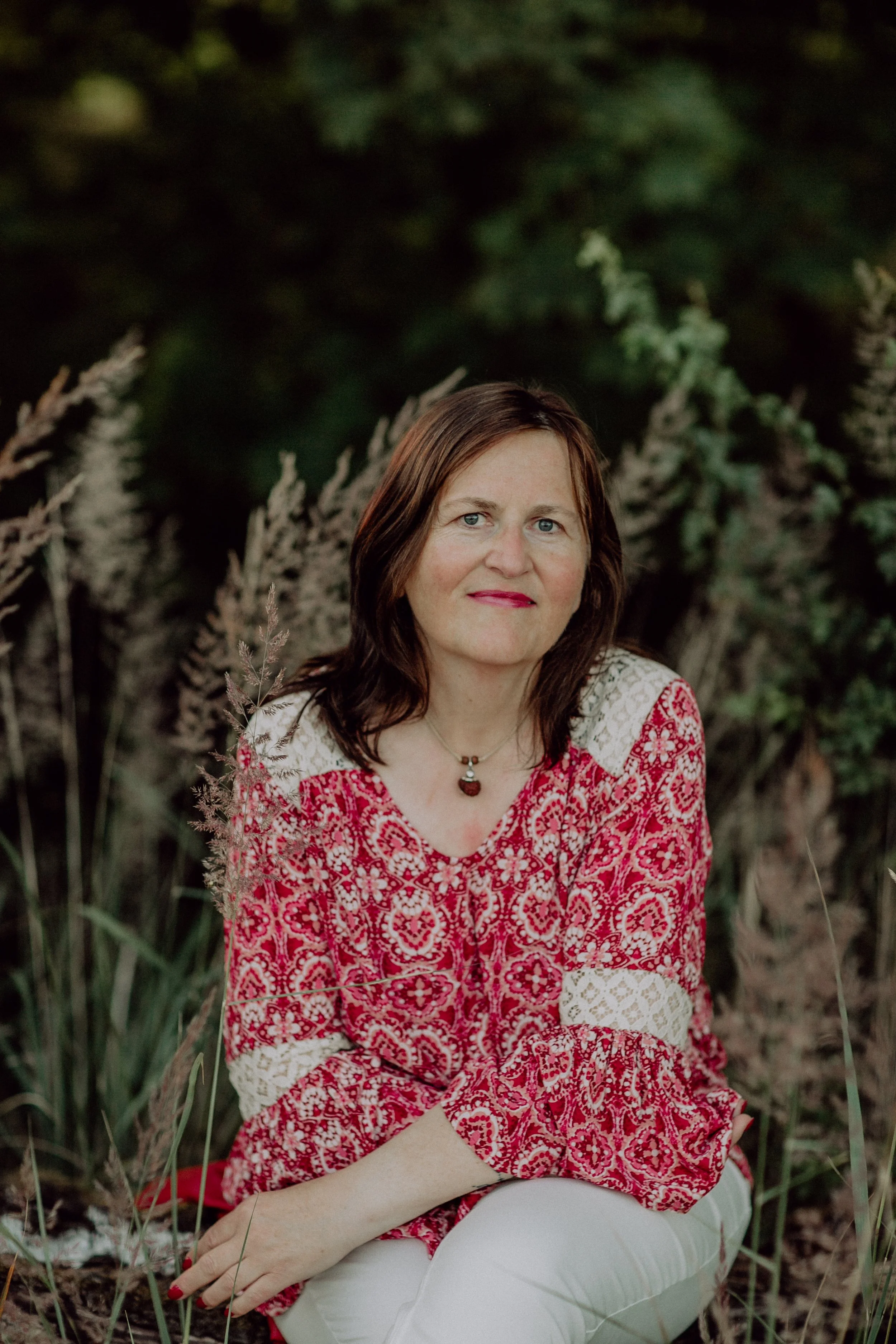 Ein Porträt einer Frau mit braunen Haaren, die in einem roten und weißen Muster-Top sitzt, im Freien mit grünen Büschen und Pflanzen im Hintergrund.