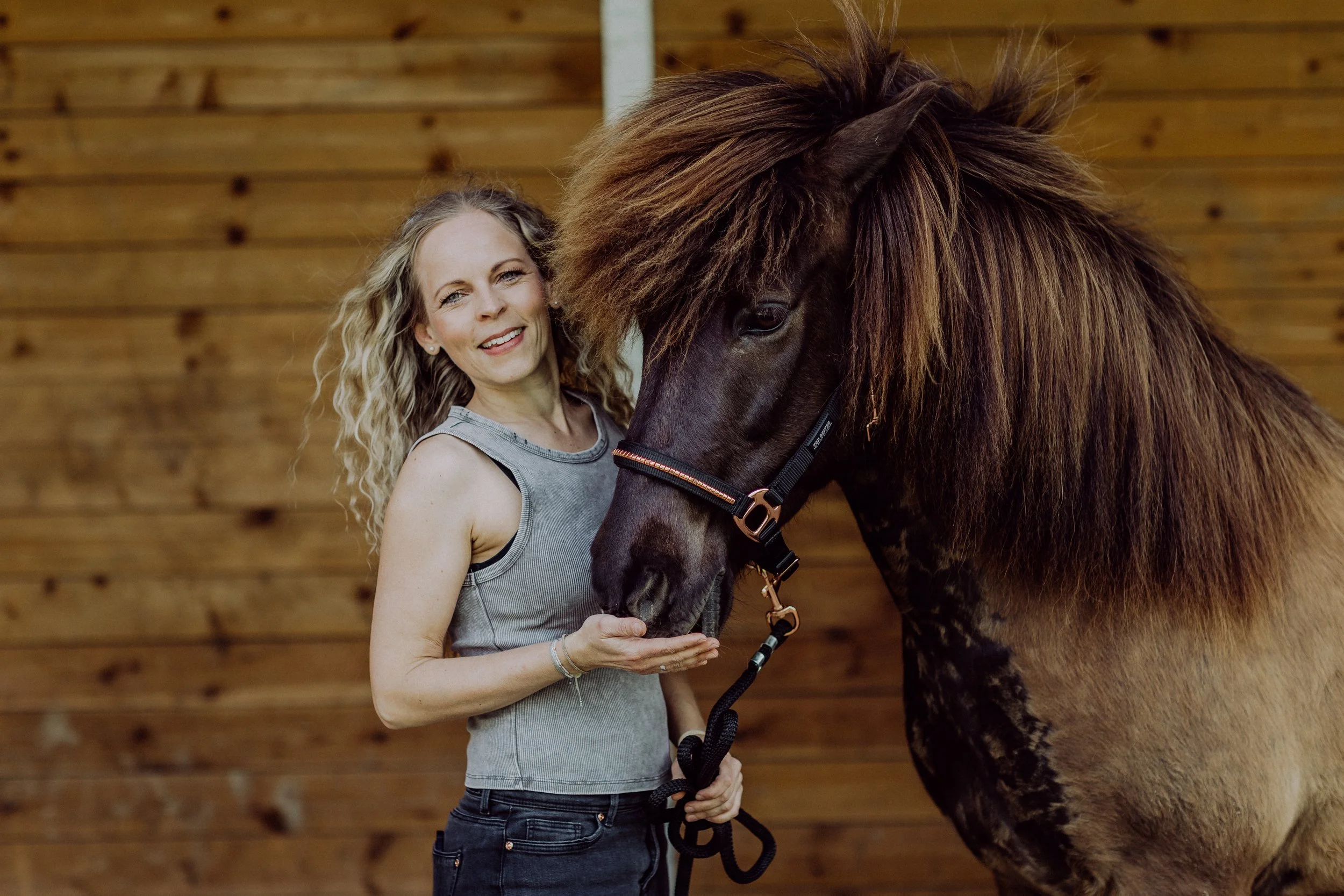 Eine Frau mit lockigem blondem Haar, trägt ein graues Top und hält die Schnur eines braunen Pferdes mit langem Haarkamm in einer Holzreithalle.