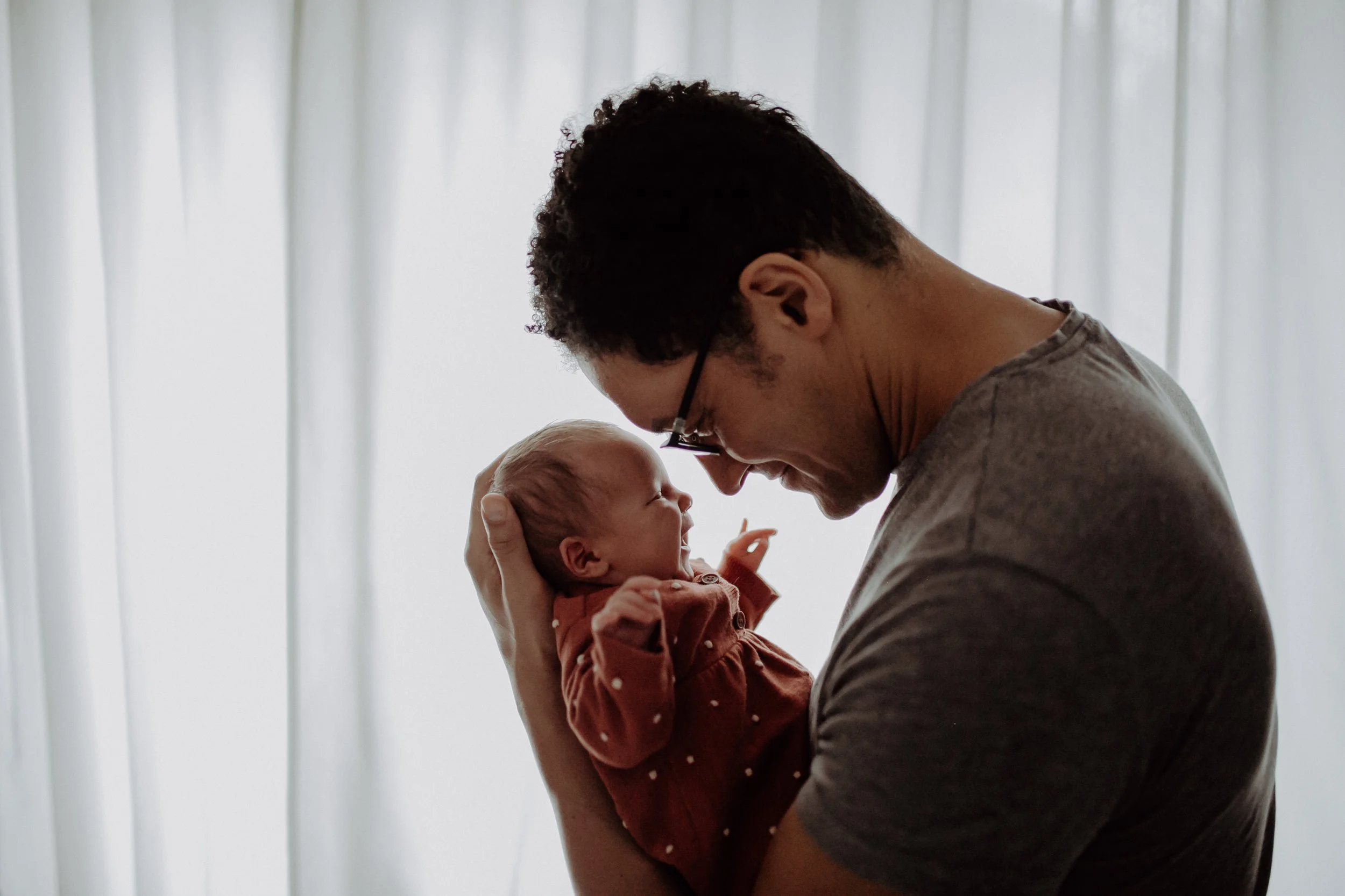 Ein Mann hält ein Baby in den Armen, beide schauen sich liebevoll an, vor einem hellen Vorhang