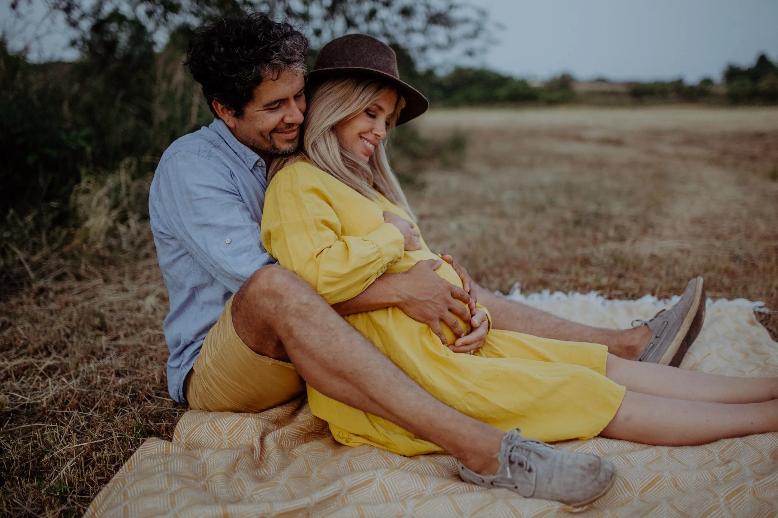 Ein Paar sitzt auf einer Decke im Freien, die schwangere Frau in einem gelben Kleid, der Mann umarmt sie und hält ihre Bauch. Es ist Abend oder Dämmerung.
