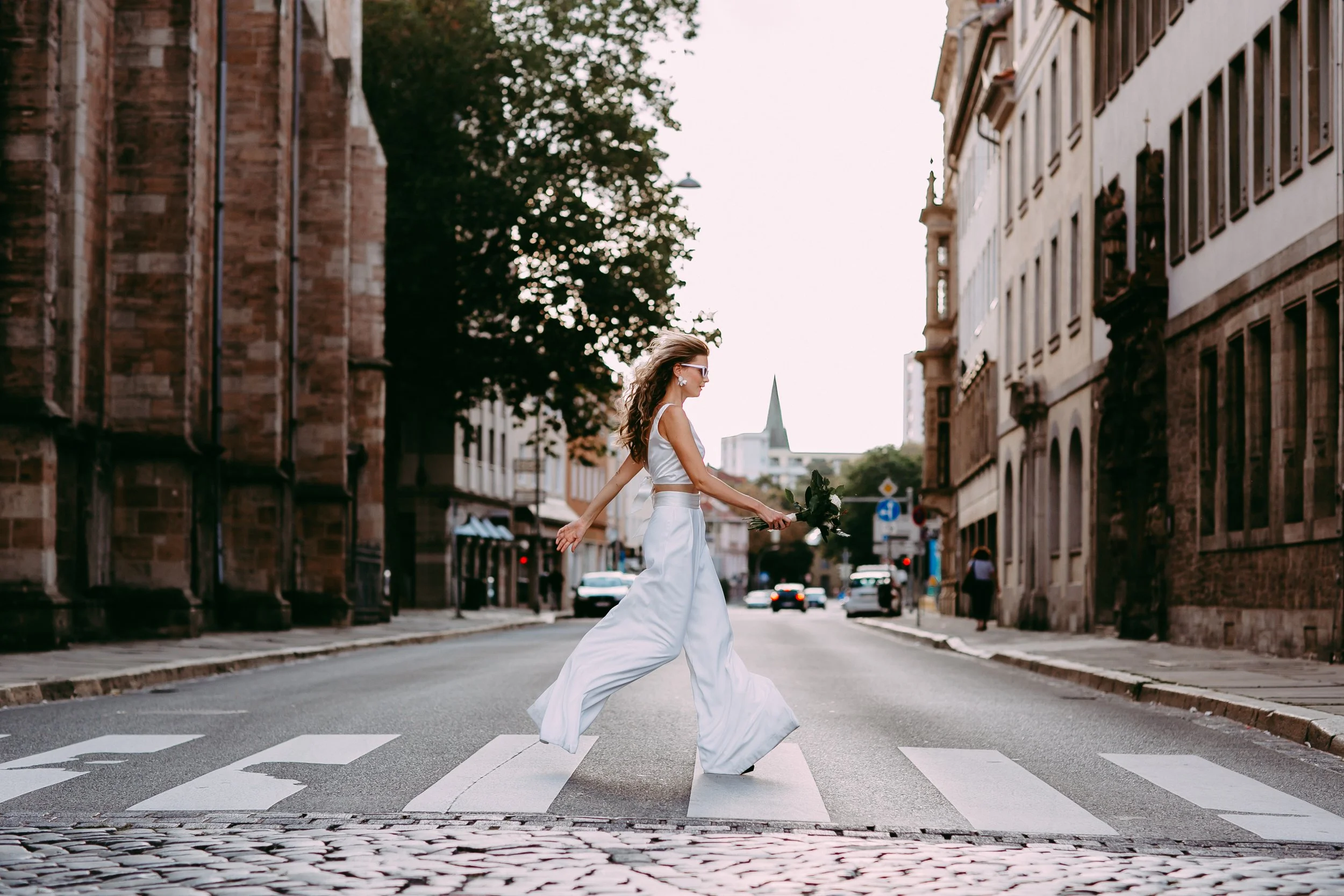 Eine Frau in einem weißen Kleid läuft auf einem Zebrastreifen in einer Stadtstraße, hält einen Blumenstrauß in der Hand und trägt Sonnenbrillen.