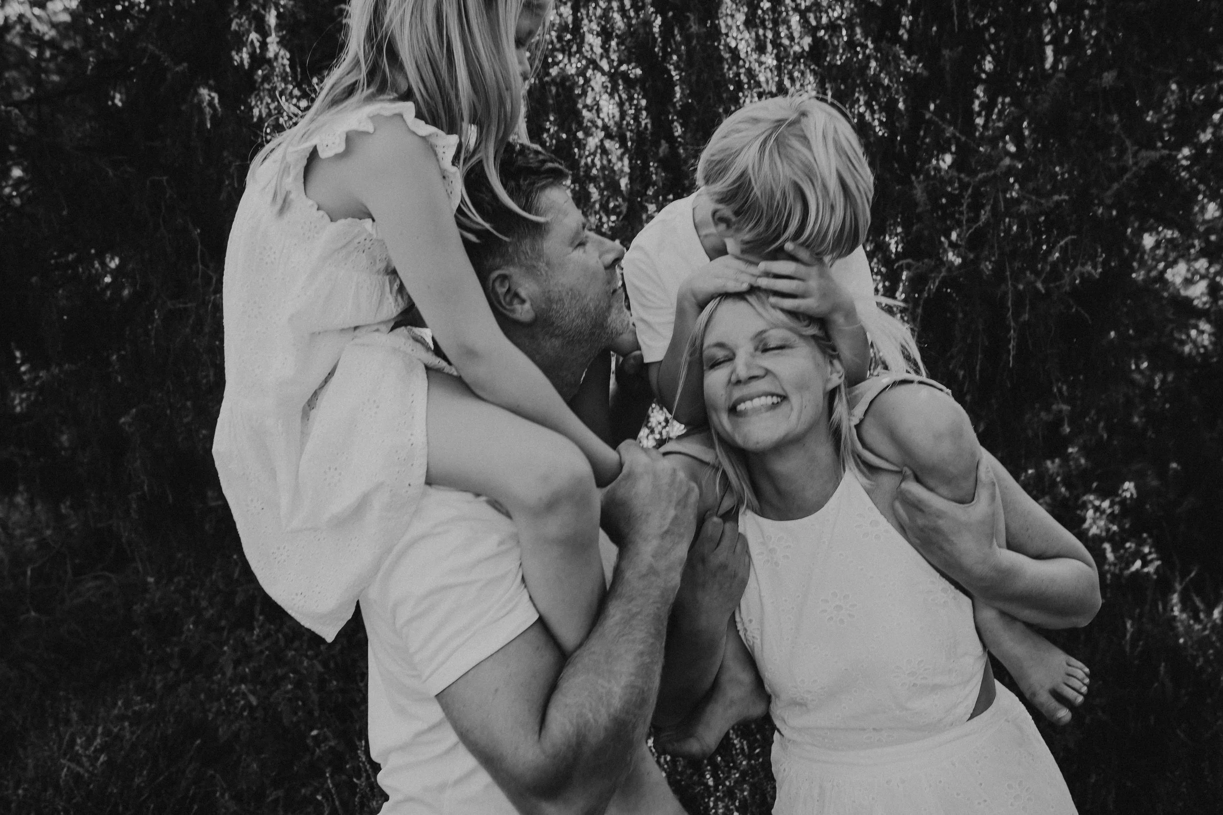 Familie mit Vater, Mutter und zwei Kindern bei einer Umarmung im Freien, lachend und umgeben von Bäumen, in Schwarz-Weiß.