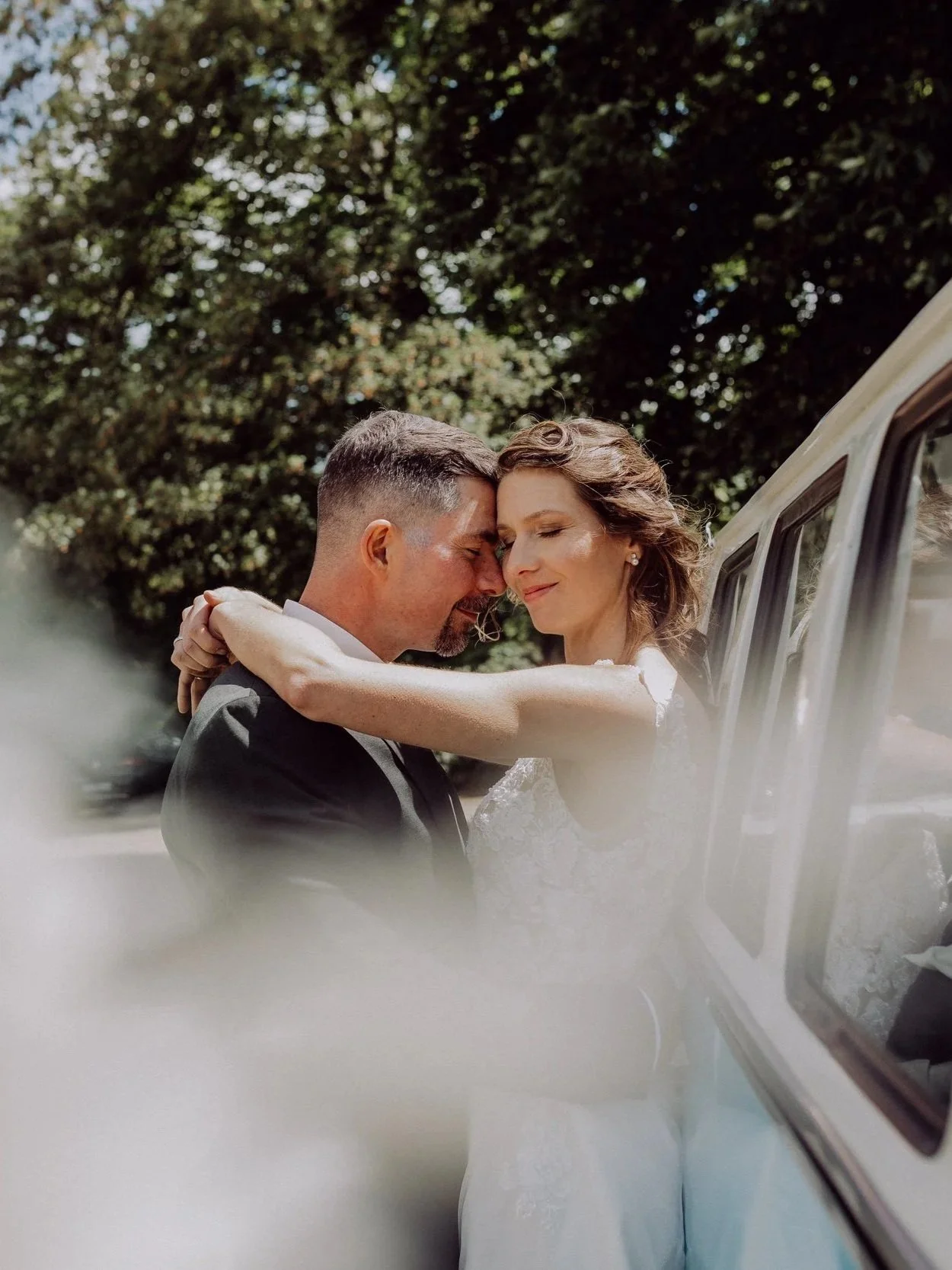 Ein Brautpaar umarmt sich vor einem Auto, bei sonnigem Wetter, mit grünen Bäumen im Hintergrund.