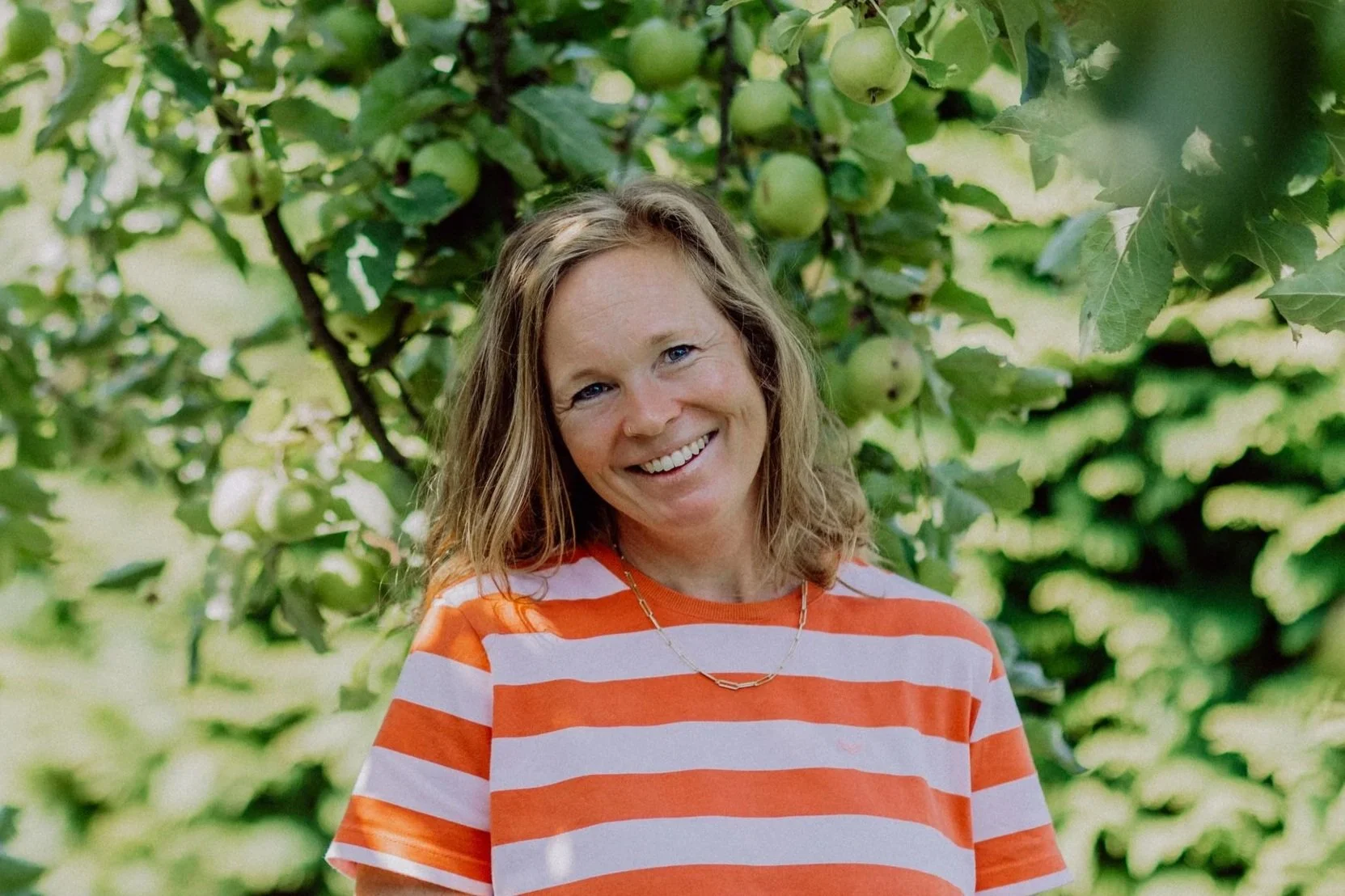 Eine lächelnde Frau mit braunen, gewellten Haaren steht vor einem Apfelbaum mit grünen Äpfeln in einem Garten.