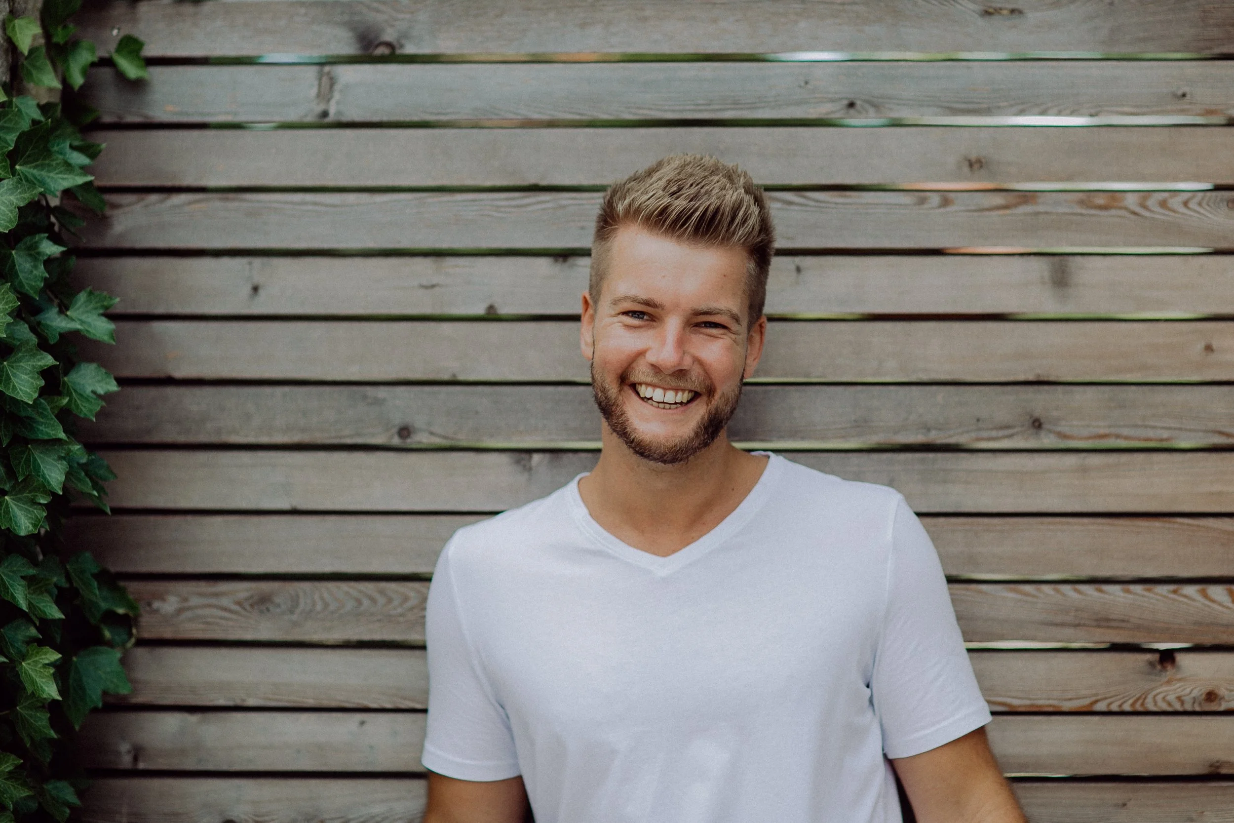 Junger Mann mit blondem Haar und Bart, lächelnd, getragen in weißes T-Shirt, vor einer Holzlattenwand, links einige grüne Pflanzen.