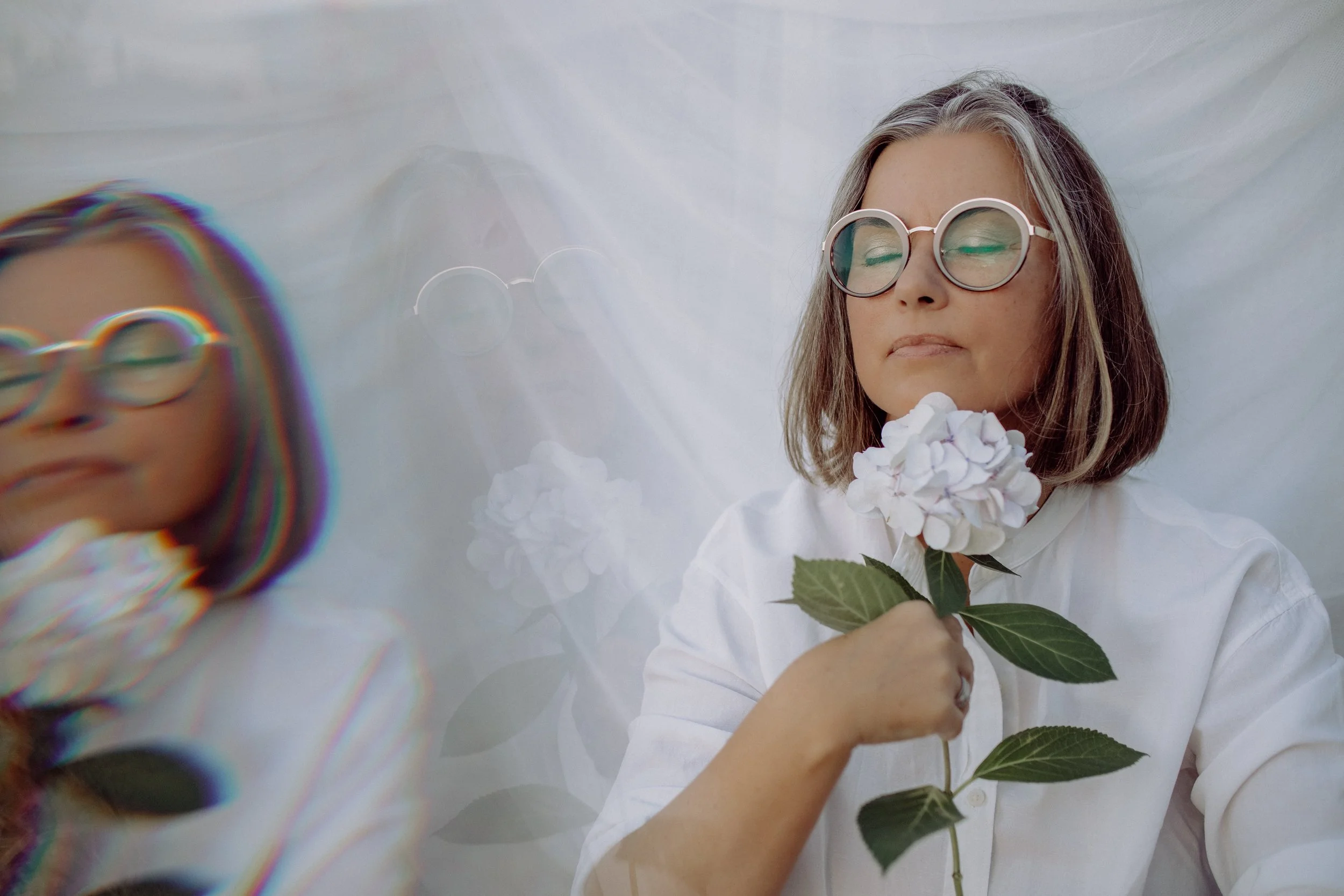 Frau mit Brille hält weiße Hortensie-Blume, blickt mit geschlossenen Augen, trägt weißes Hemd, im Hintergrund weiße Stoffwand, lila-blauer Schatten auf der linken Seite.