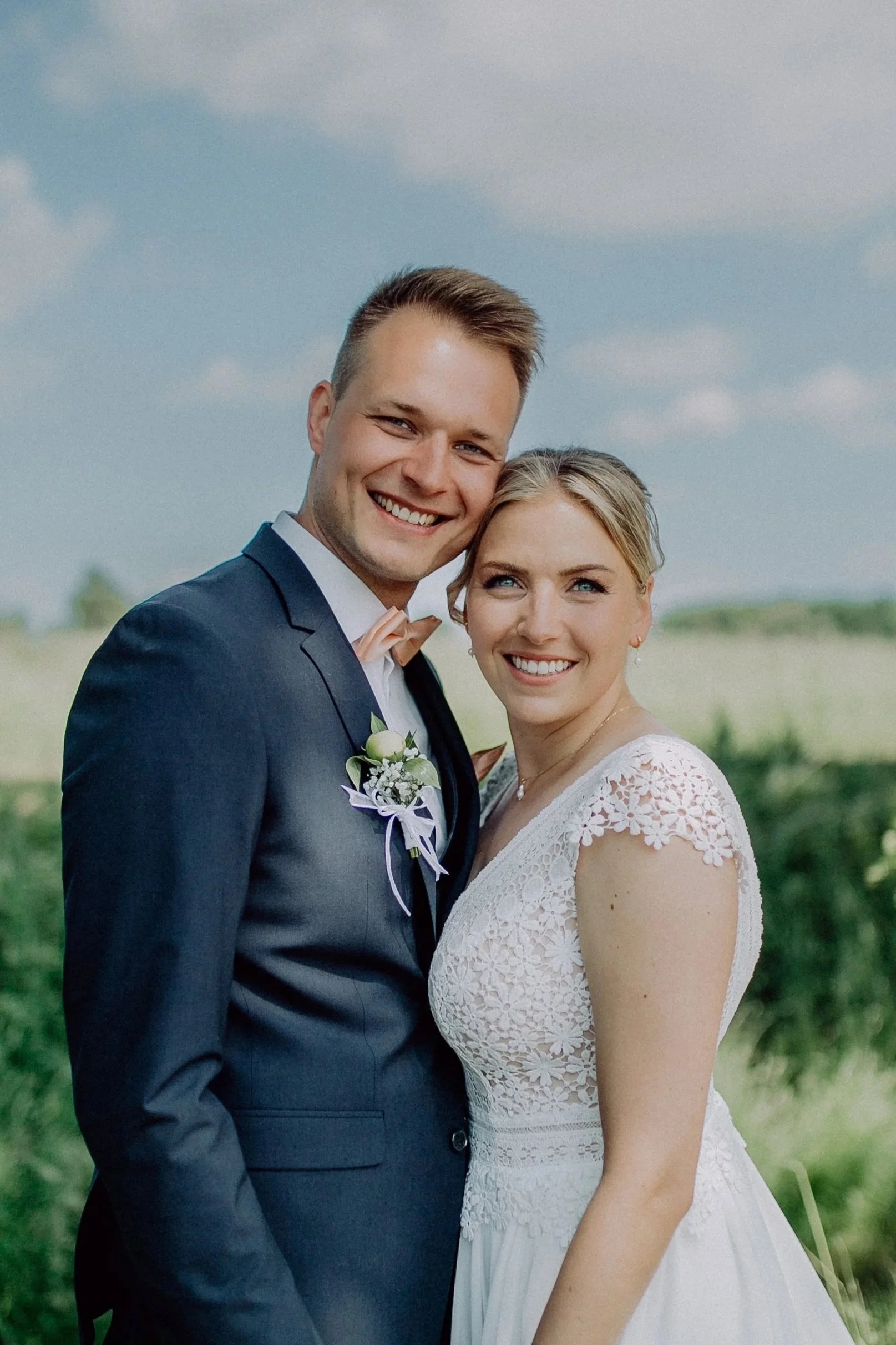 Ein glücklich wirkendes Brautpaar im Freien bei Sonnenlicht, der Mann trägt einen dunkelblauen Anzug mit Fliege, die Frau trägt ein weißes Spitzenkleid, beide lächeln in die Kamera.