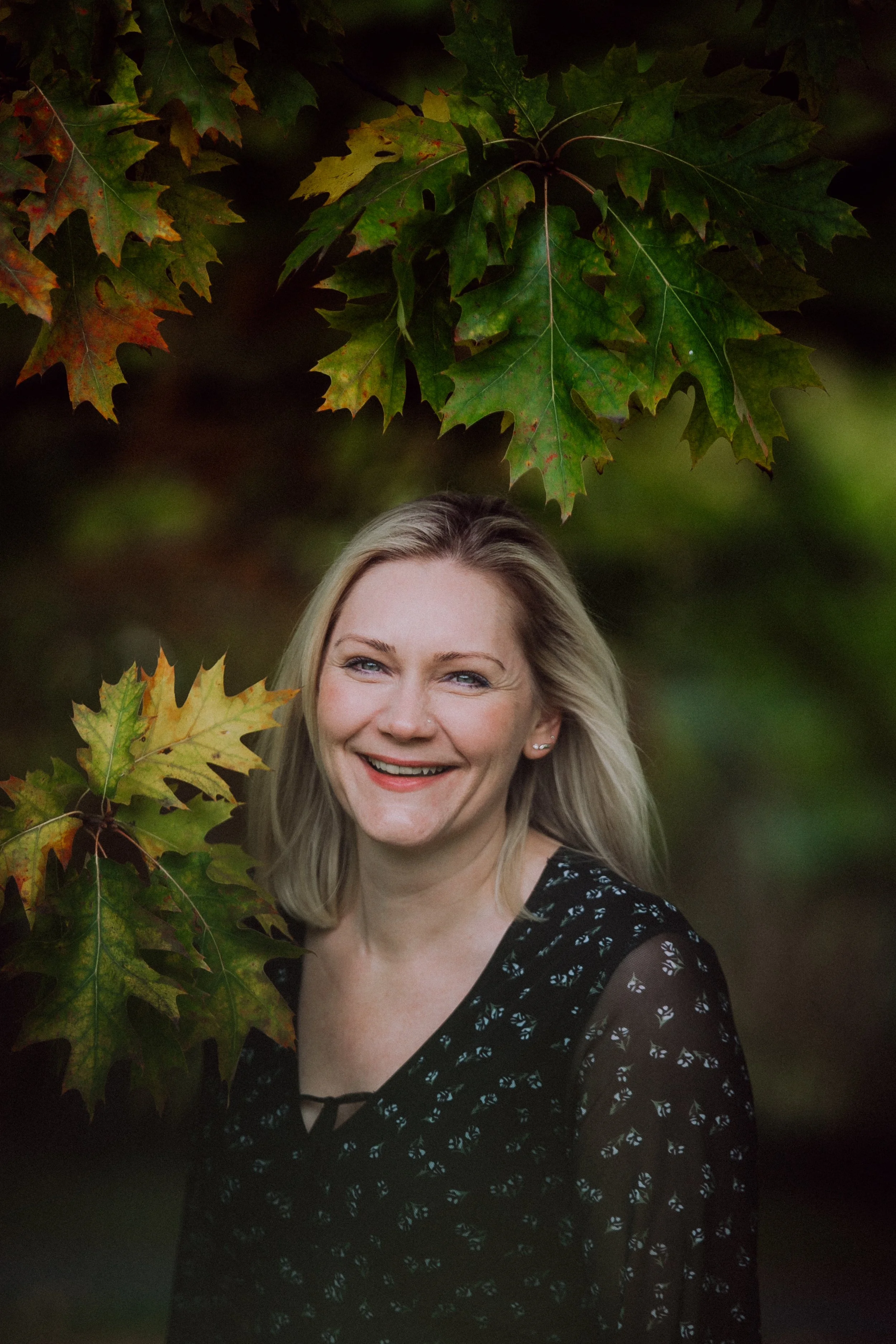 Lächelnde Frau mit blonden Haaren vor herbstlichen Blättern.