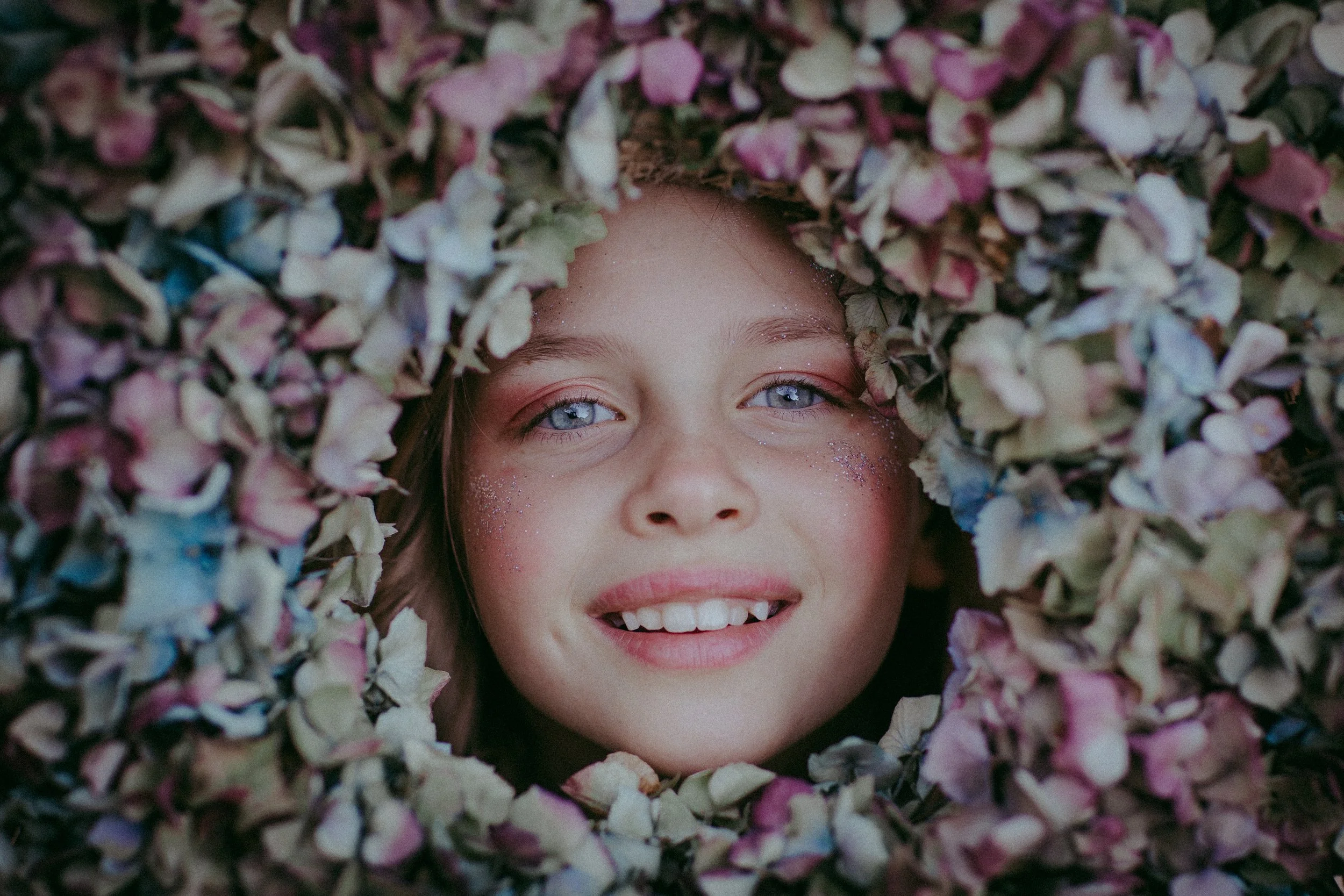 Nahaufnahme eines lachenden Mädchens mit blauen Augen, umgeben von getrockneten Blumen.