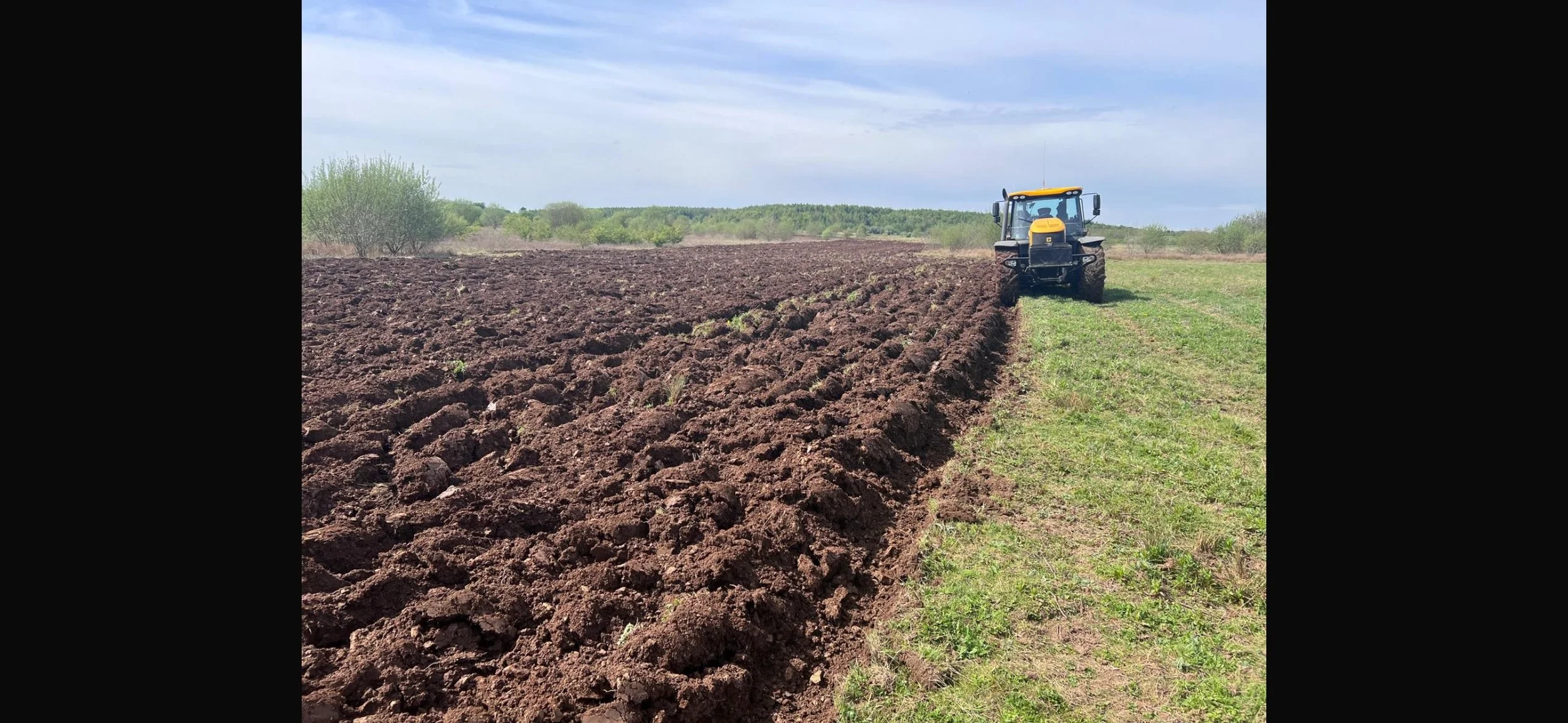 A tractor working on a field with freshly tilled dark brown soil, with green grass on the right side and trees in the background under a cloudy sky.
