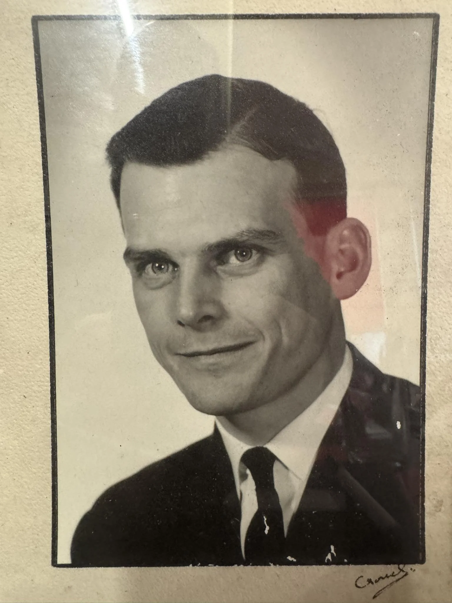 Black and white portrait photograph of a man in a suit and tie, with neatly combed hair, smiling slightly, signed at the bottom right corner.