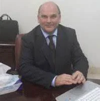 A man in a suit sitting at a desk with his hands clasped, smiling, in an office setting.