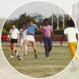 Four young people playing soccer on a grassy field, with trees and a sports fence in the background.