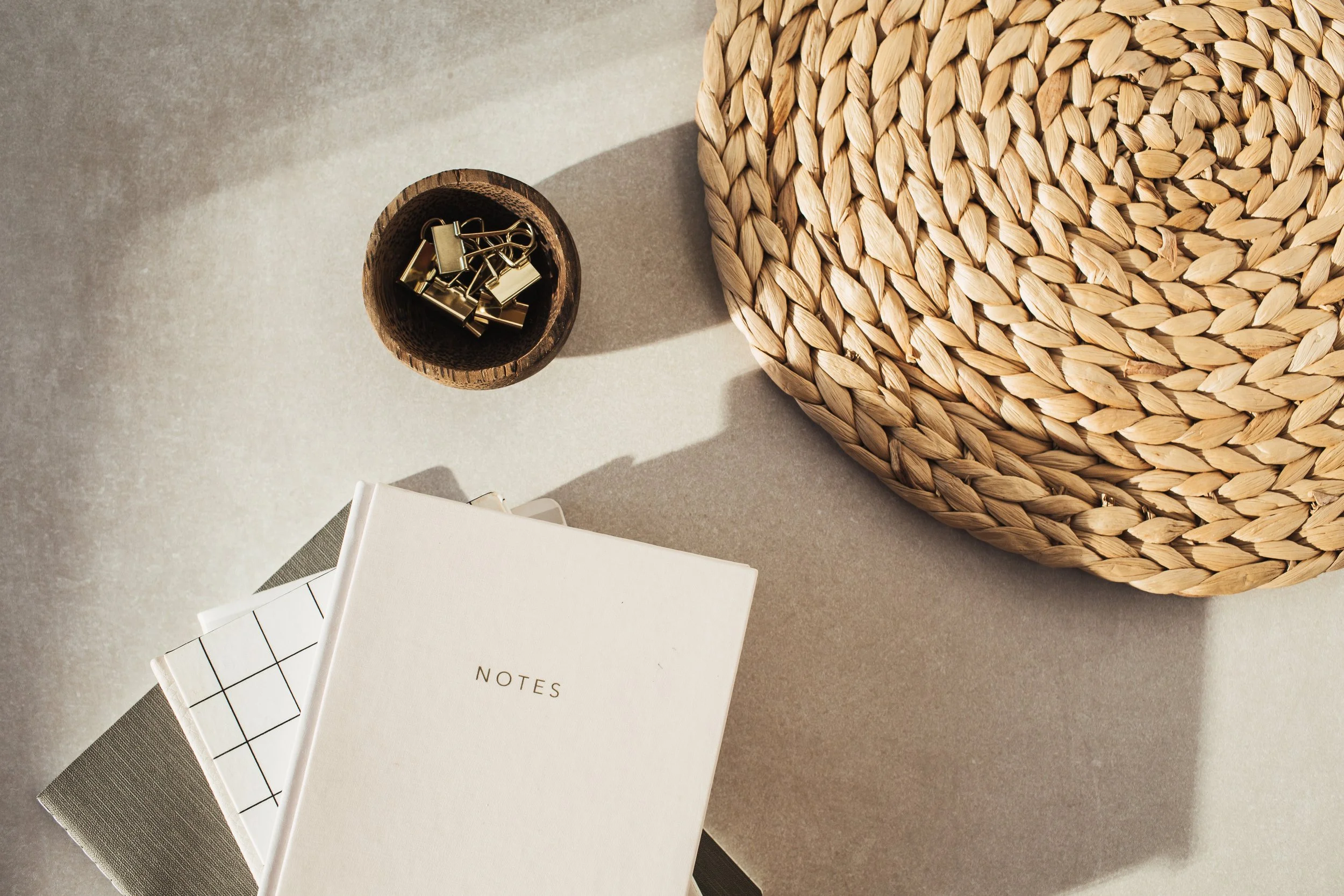 Top-down view of a desk with a woven placemat, a closed notebook labeled 'NOTES', a small stack of notepads, and a small wooden bowl containing gold binder clips, with sunlight casting shadows.