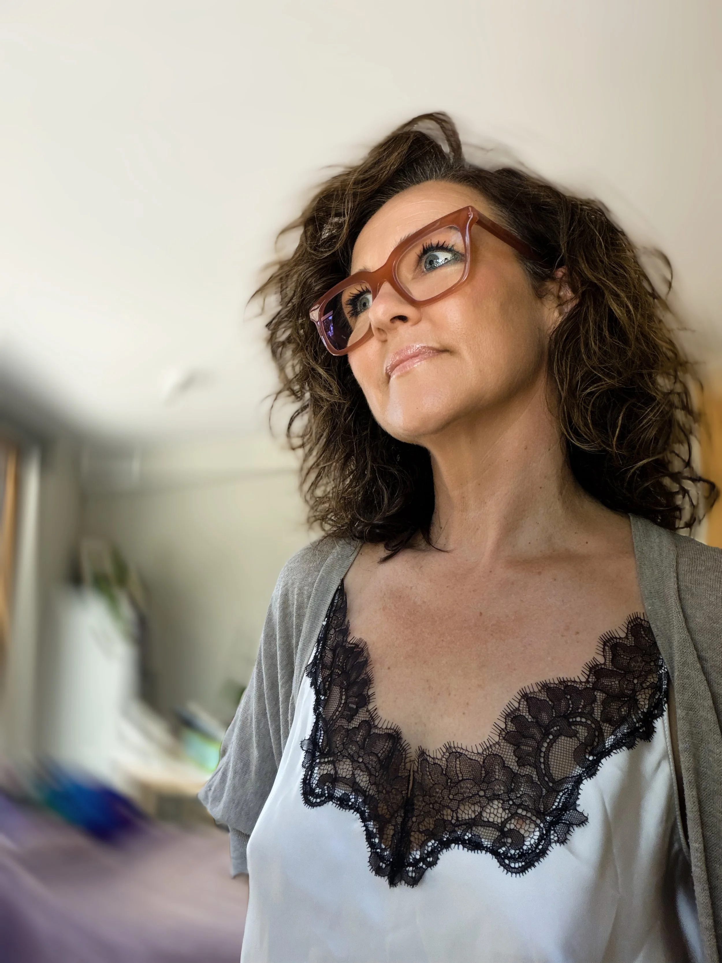 A woman with curly brown hair wearing red glasses and a camisole with black lace detail, looking upwards.