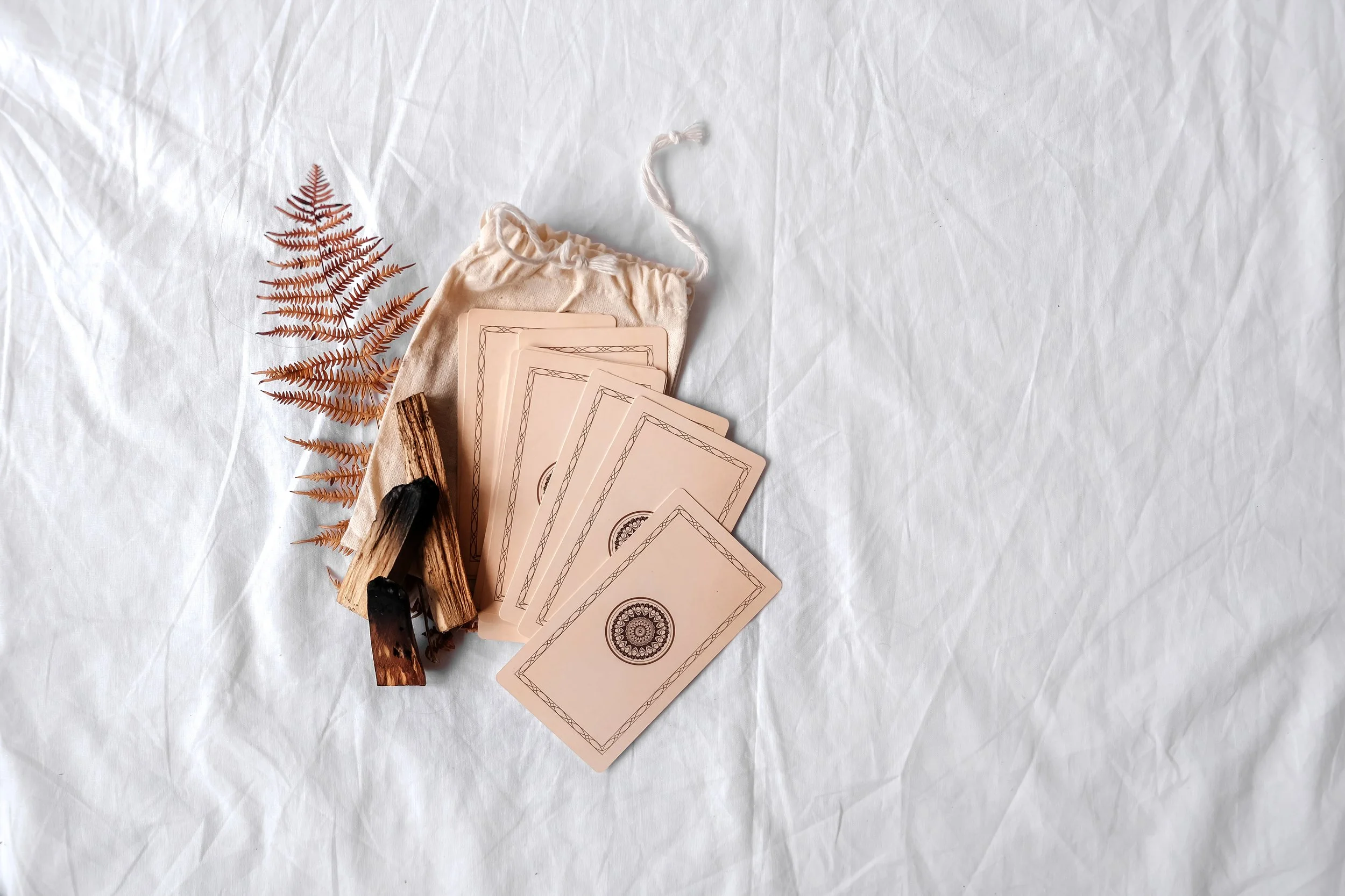 A set of tarot cards, dried fern leaf, driftwood, and a cloth pouch laid on crumpled white paper.