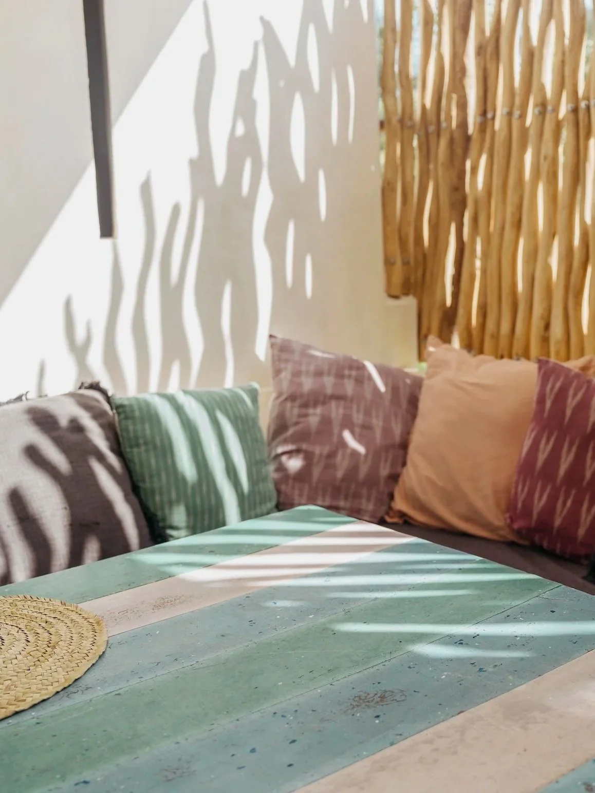 Outdoor seating area with multicolored cushions, a recycled plastic table in pastel colours, and a decorative wooden privacy screen casting shadow patterns on the wall and table.