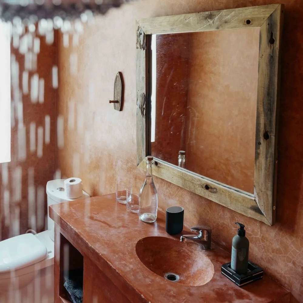 A rustic bathroom with a reddish-orange textured wall, featuring a mirror with a wooden frame above a matching reddish-orange countertop with an oval sink. 
