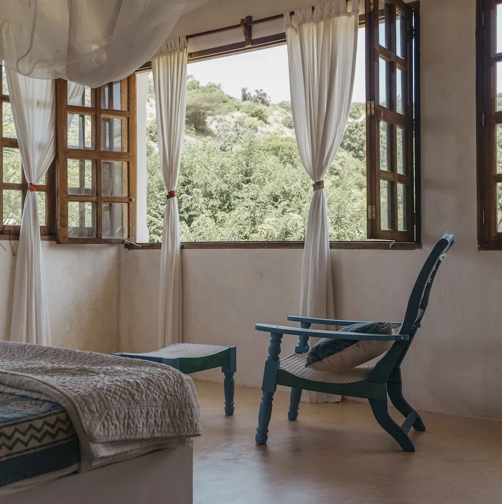 A bedroom with open wooden windows, white curtains, a green recycled plastic chair with a cushion, and a view of green trees and hills outside.