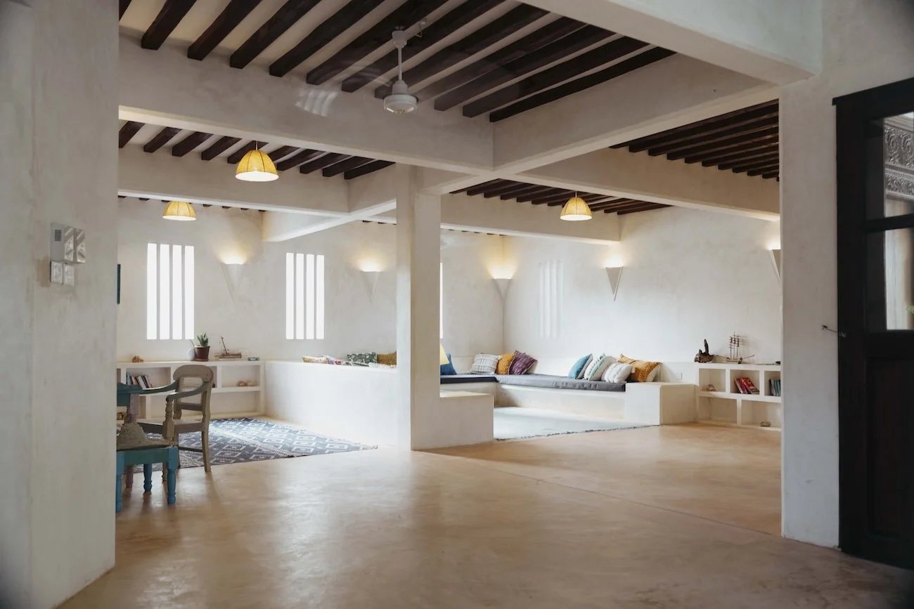 Spacious living room with white walls, dark wooden ceiling beams, built-in seating with cushions, small shelves with books and decorative items, and soft lighting.