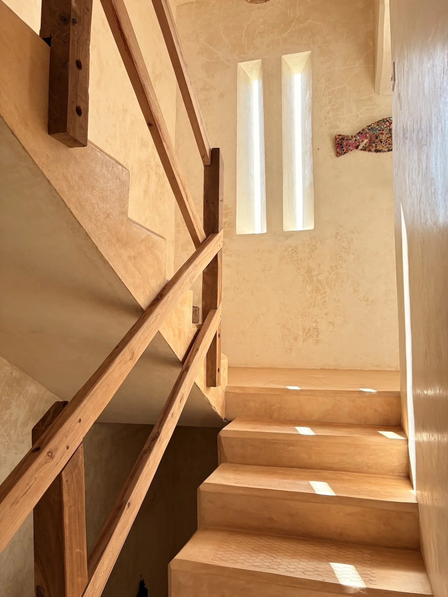 Wooden staircase with a railing, leading upwards with sunlight streaming through narrow windows on a textured yellow wall.
