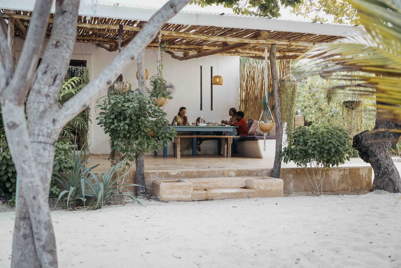 People dining on a covered patio with greenery and sand in the foreground.