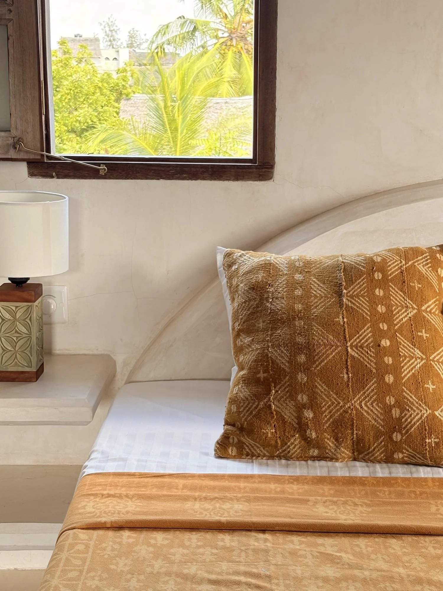 A lushly made up bed with soft linens and African prints, a window behind looks out to palm trees.
