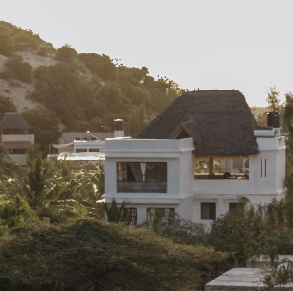 A white house with a makuti roof nestled in sand dunes, surrounded by trees and other buildings, during sunset.