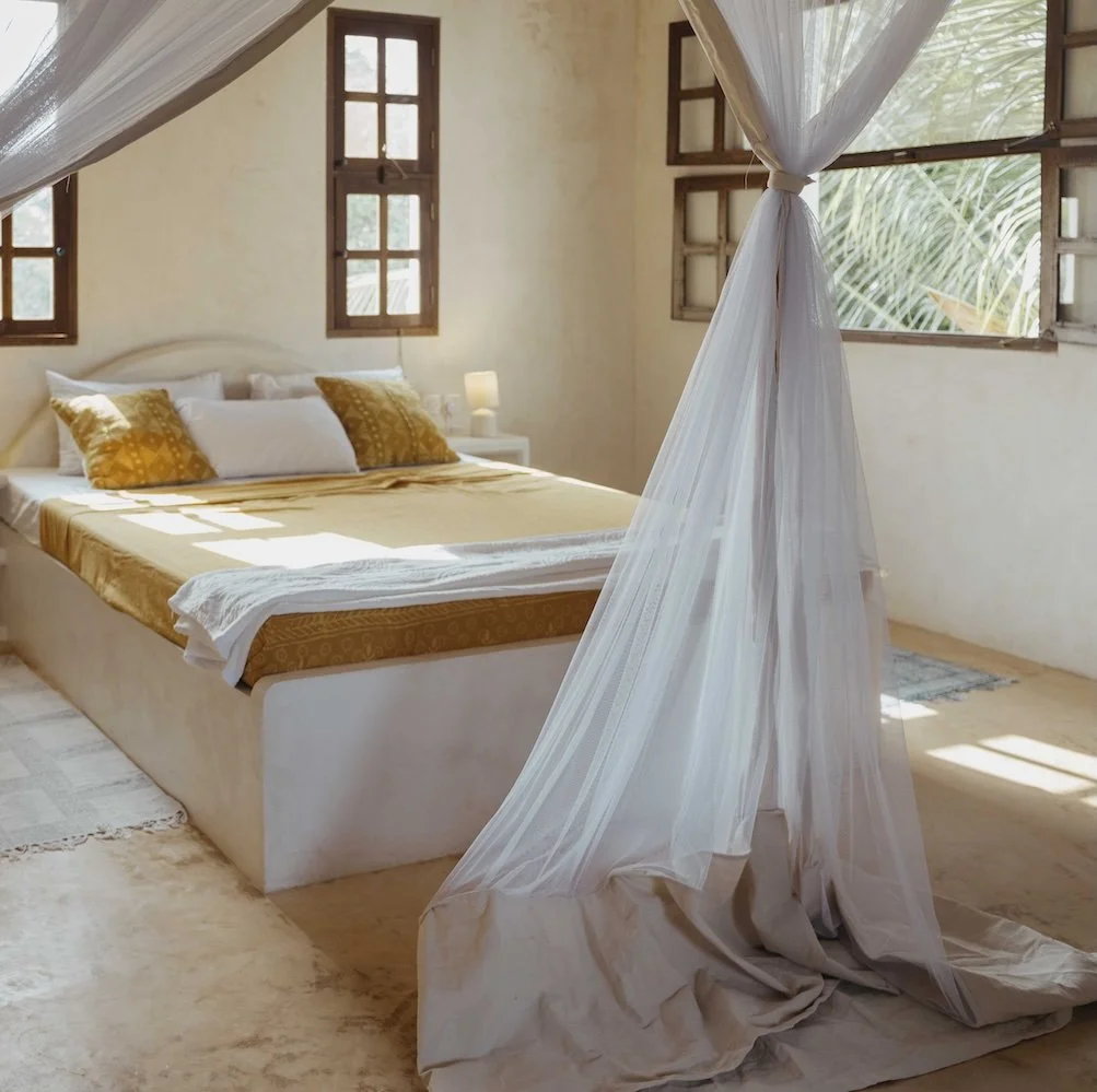 A bedroom with a bed featuring yellow and white bedding, surrounded by windows with wooden frames, and with a mosquito net.