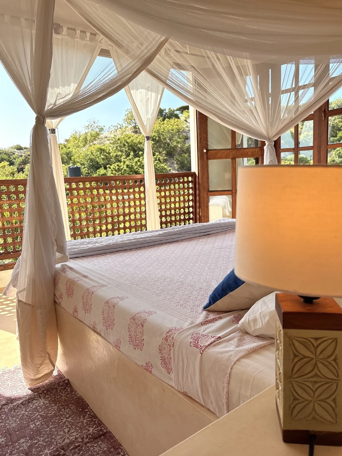 Sunlit top bedroom with a mosquito net, white curtains, and a wooden juliet balcony looking out to the Shela dunes.