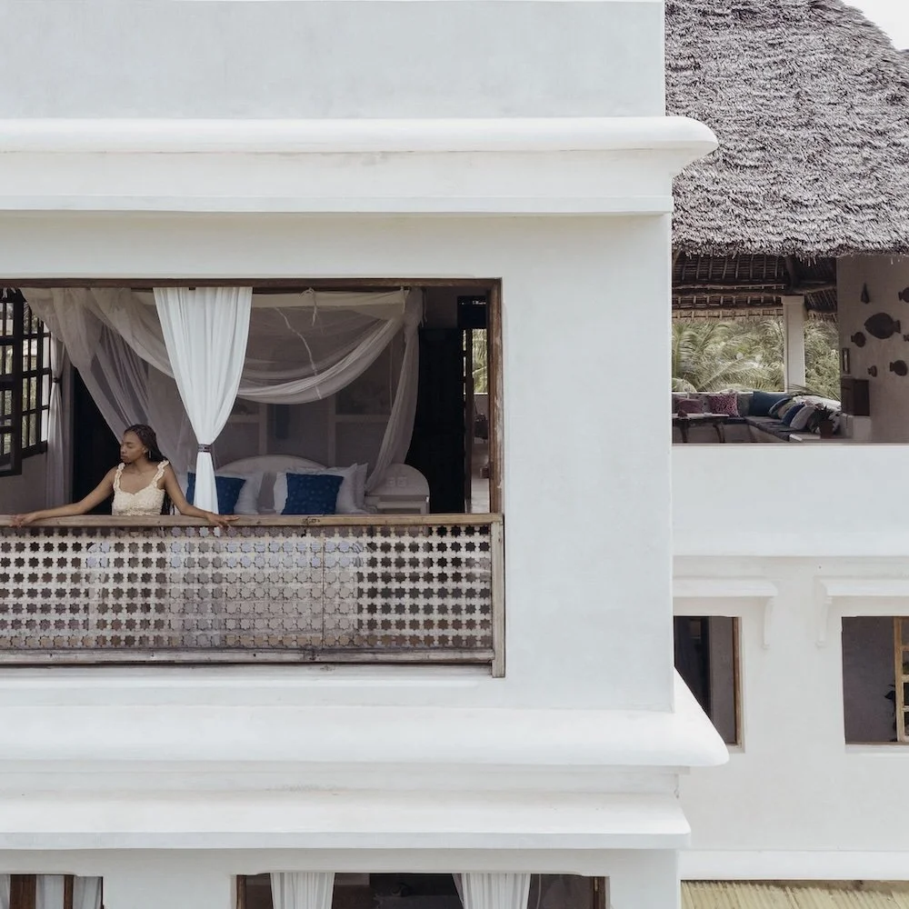 Woman relaxing on a balcony in a white dress beneath white mosquito netting, overlooking a room with beds, in a tropical resort setting.