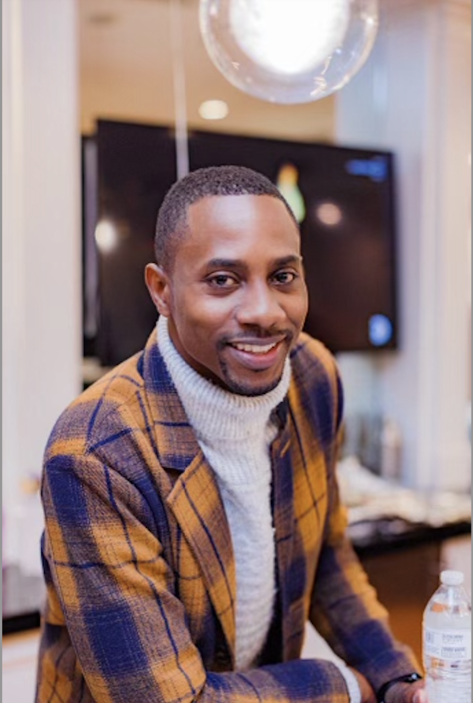 A smiling man with short hair, wearing a tan and purple plaid blazer and a white turtleneck sweater, sitting at a table indoors.