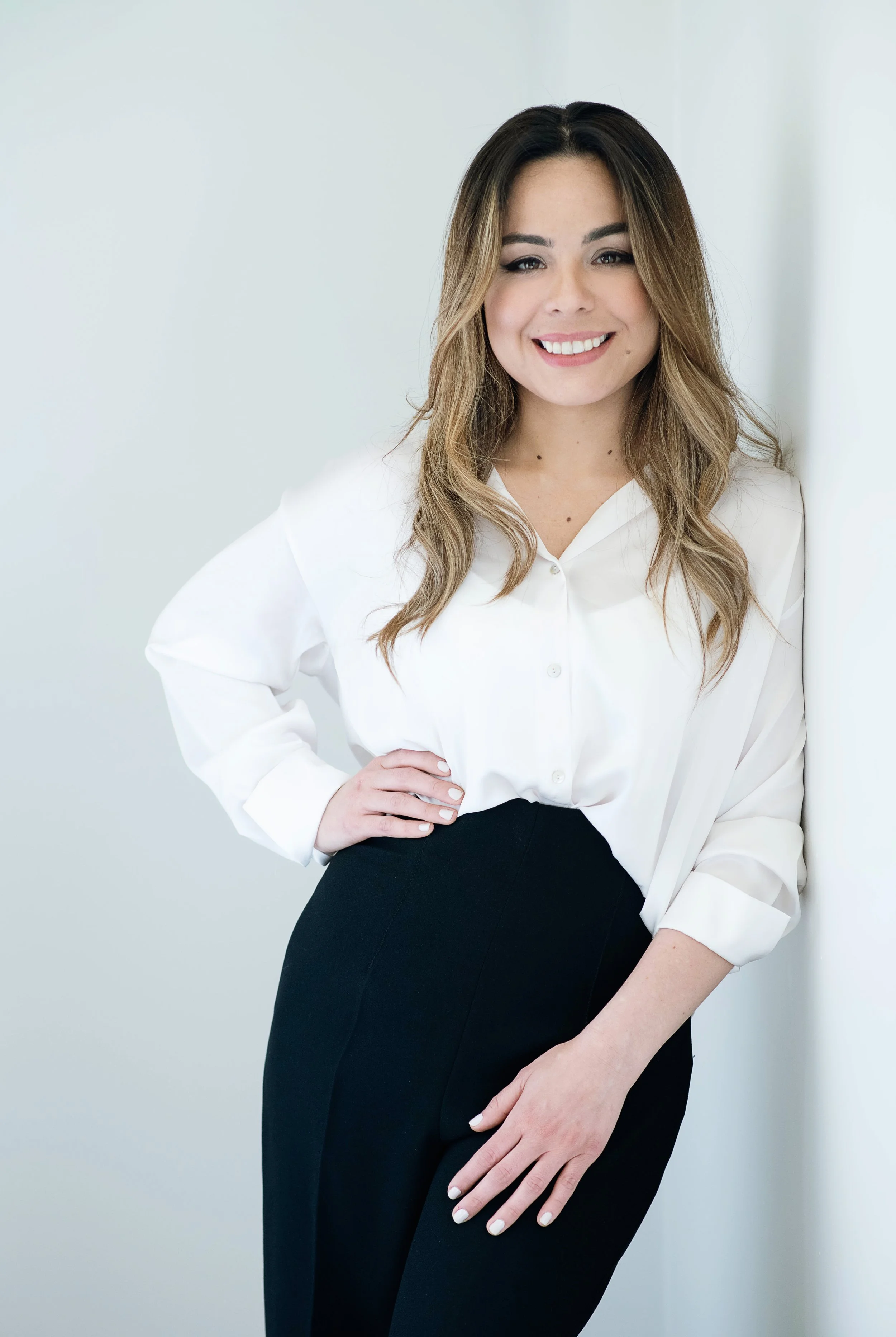 A young woman with long wavy brown hair, wearing a white blouse and black pants, smiling and leaning against a white wall.