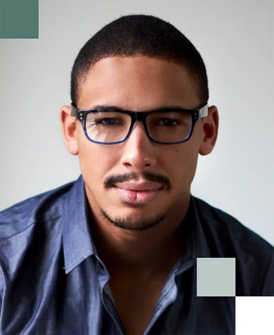 Close-up of a young man with short dark hair, wearing glasses and a dark blue shirt, looking directly at the camera against a plain background.